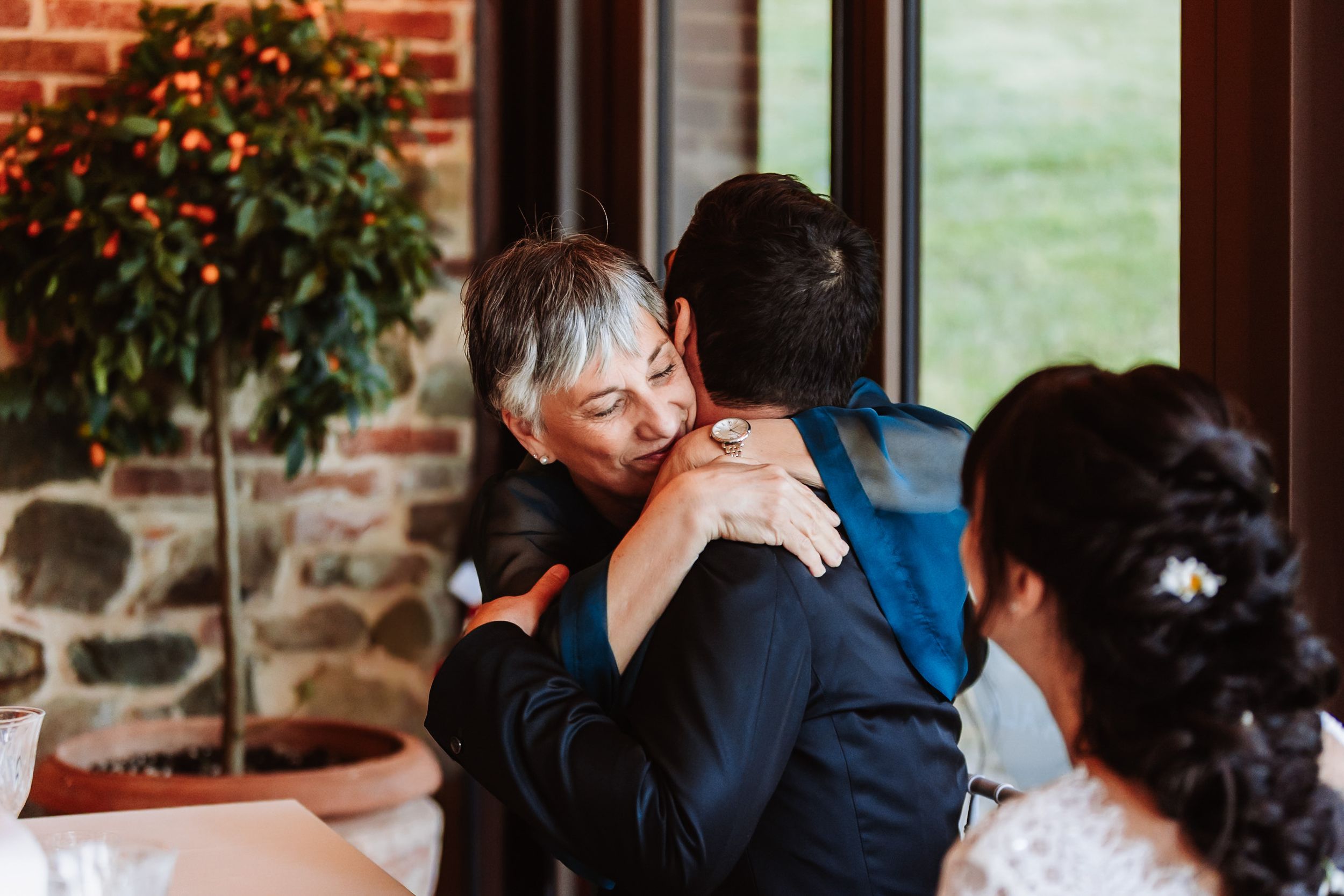 Emotional wedding moment as older woman with short gray hair hugs groom in blue suit indoors.