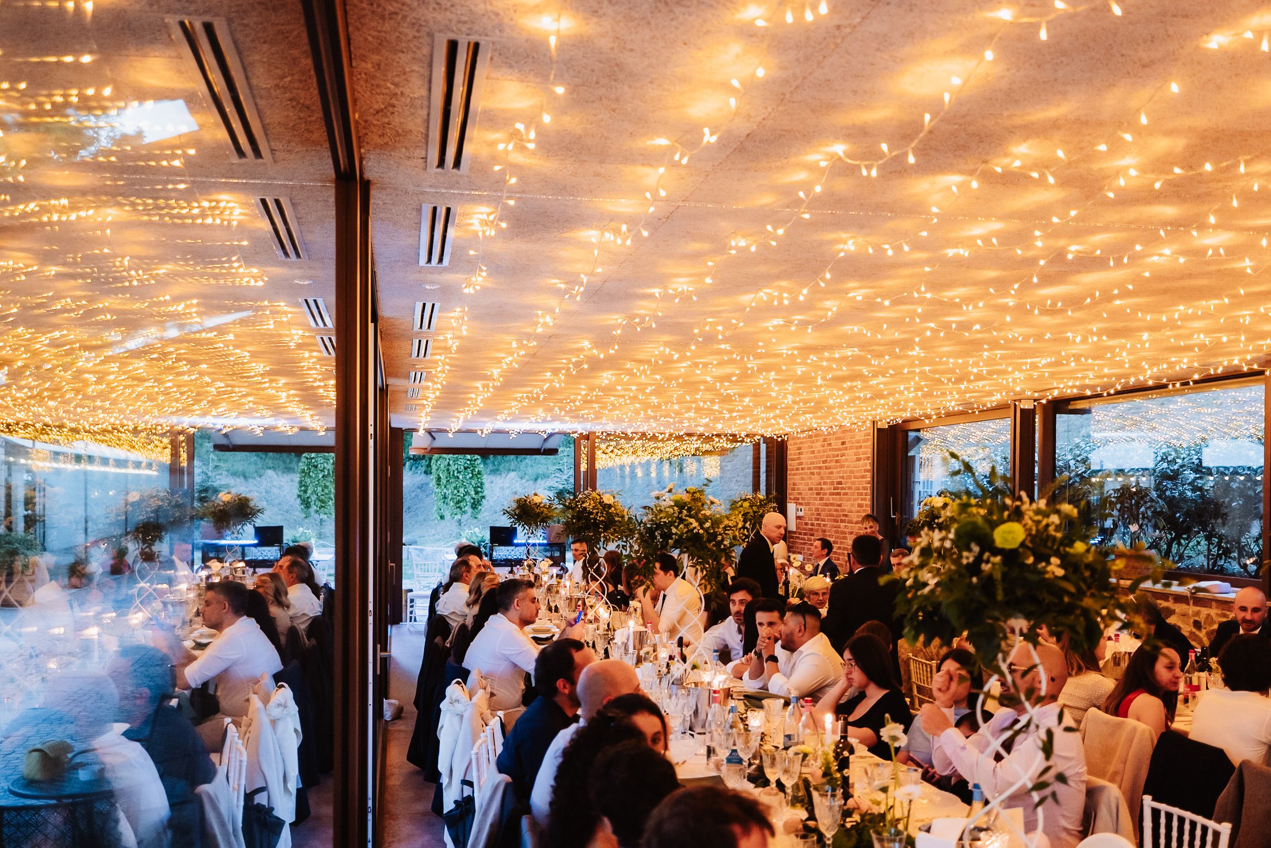 Elegant wedding reception venue with fairy lights ceiling, guests dining at decorated tables with lush greenery centerpieces.