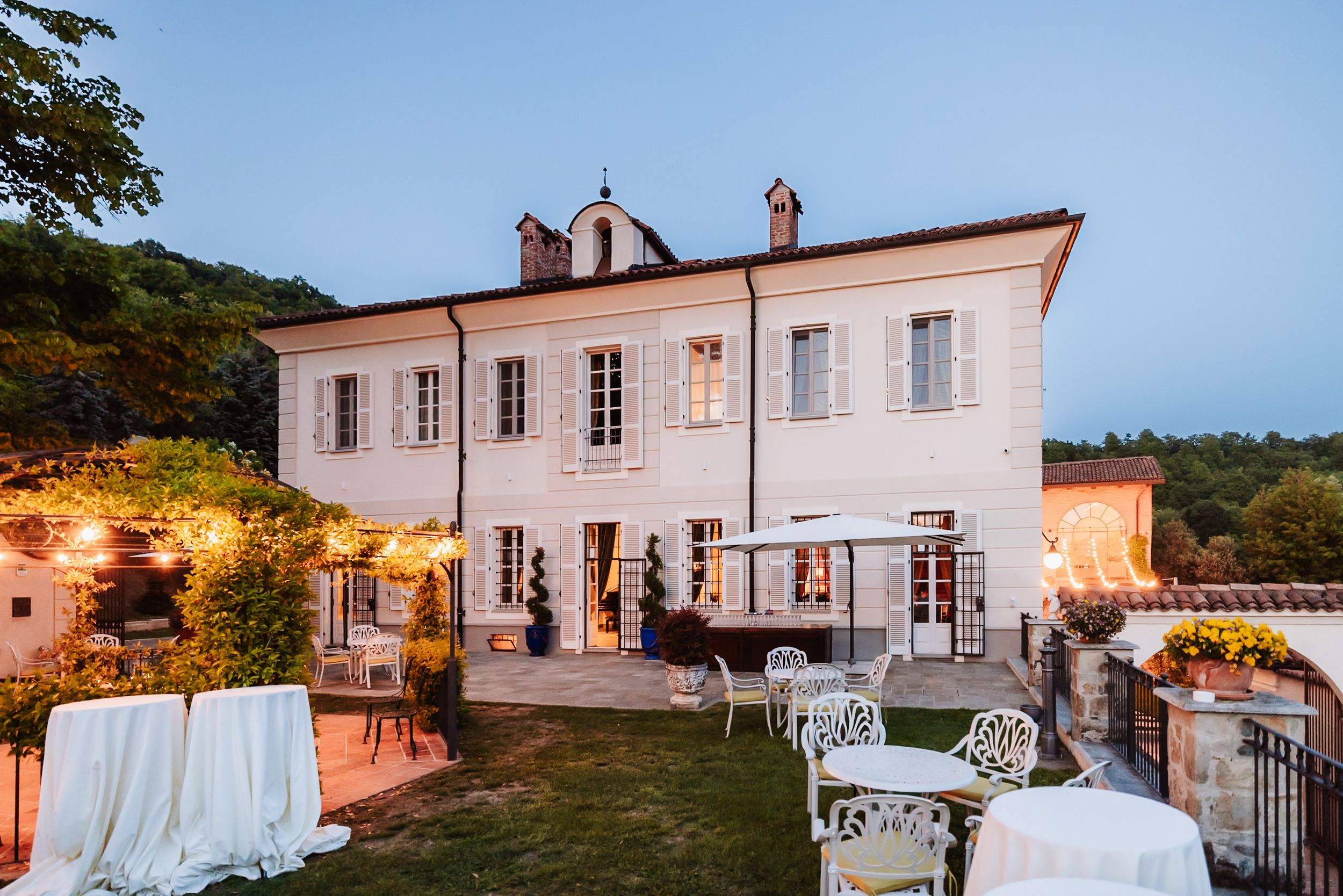 Elegant white French villa with outdoor event setup, string lights, and manicured garden at dusk.