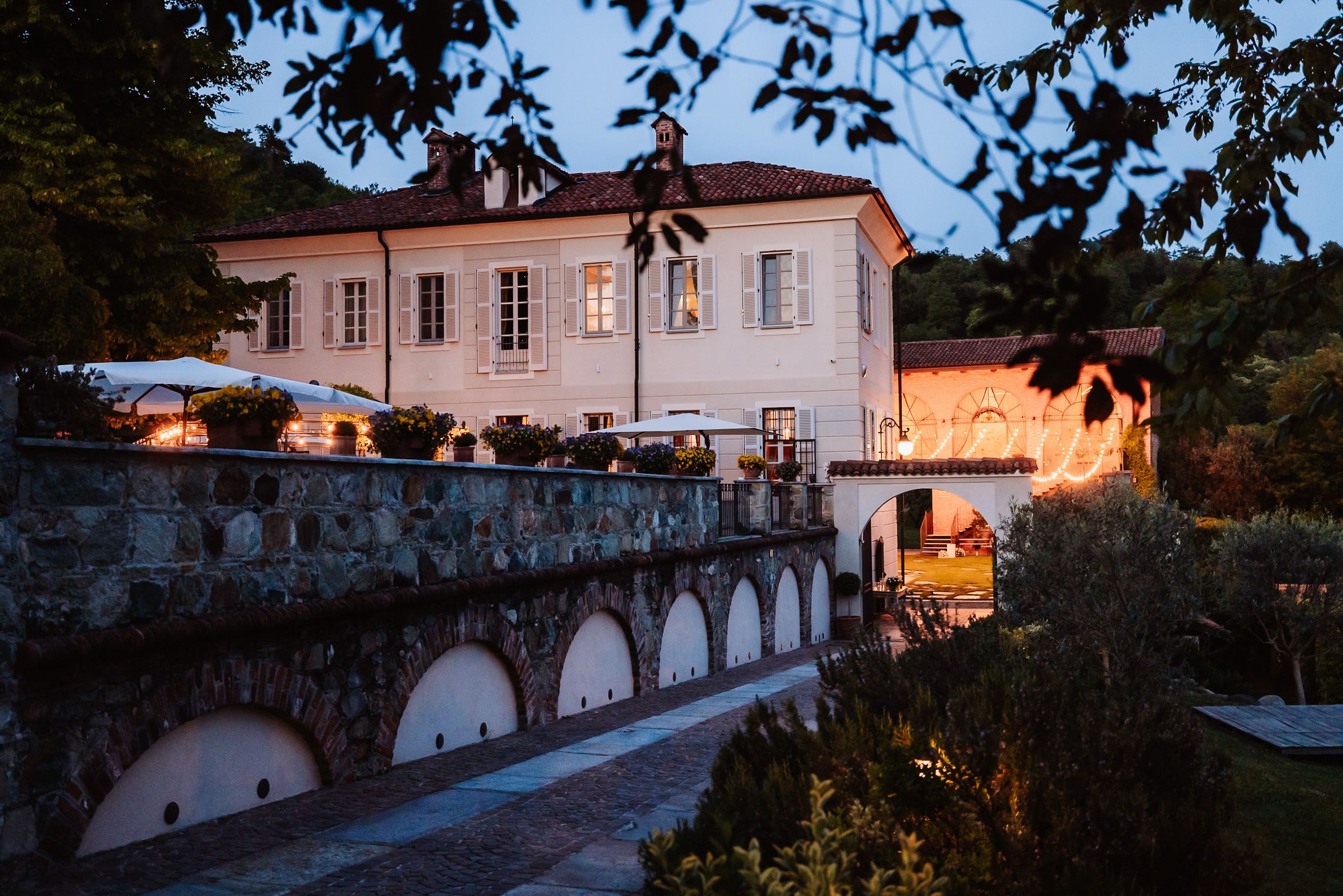 Elegant Italian villa illuminated at dusk with stone arched facade, lush gardens, and warm glowing windows.