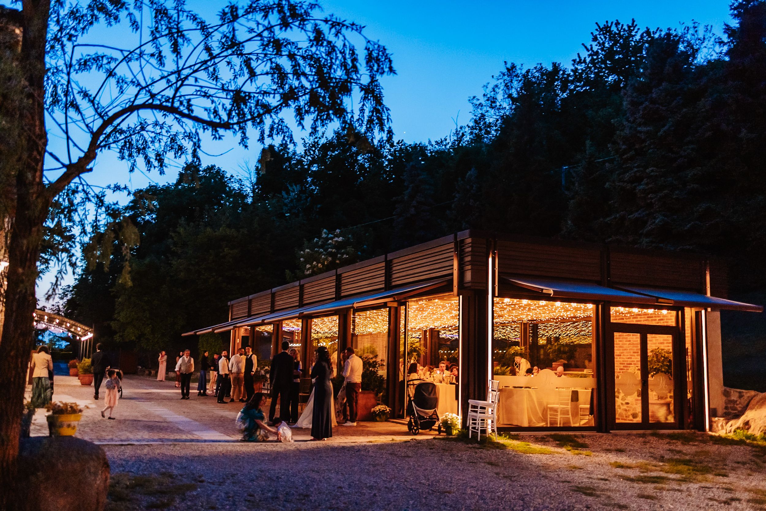 Illuminated outdoor venue at dusk surrounded by trees with guests gathered outside during an evening event.