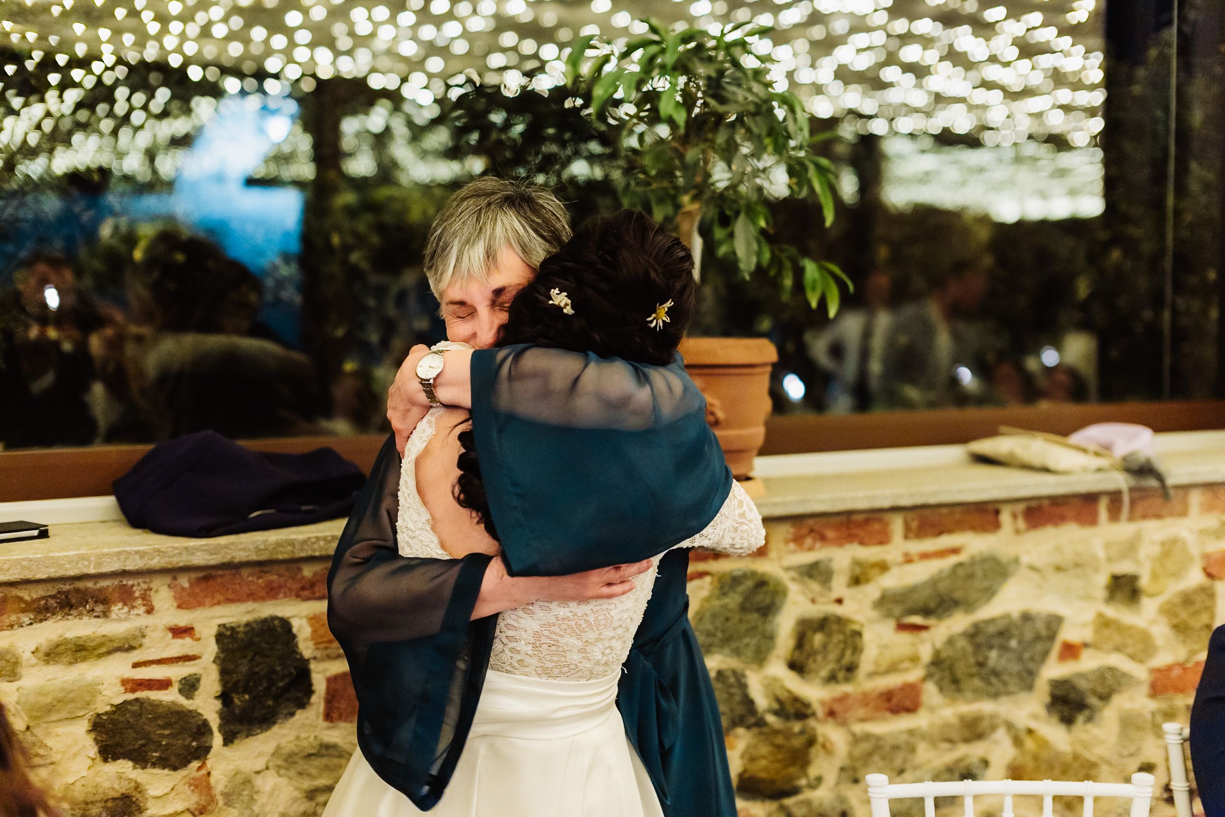 Two women share an emotional hug at a wedding reception, one in a white bridal gown, set against a rustic stone wall backdrop.