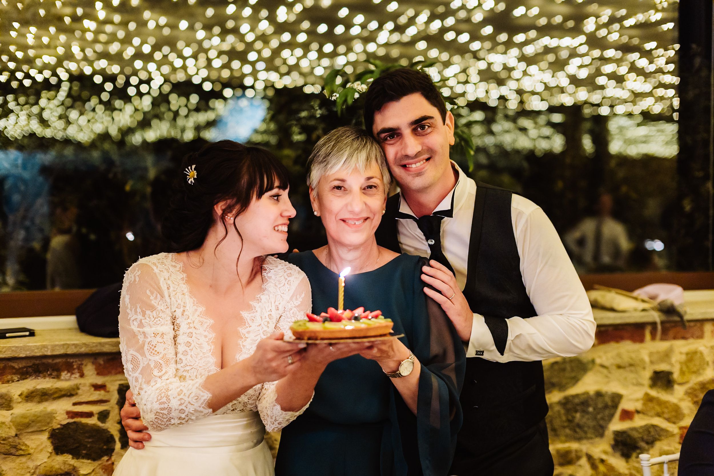 Bride and groom holding a birthday cake with candle alongside older woman at wedding reception.