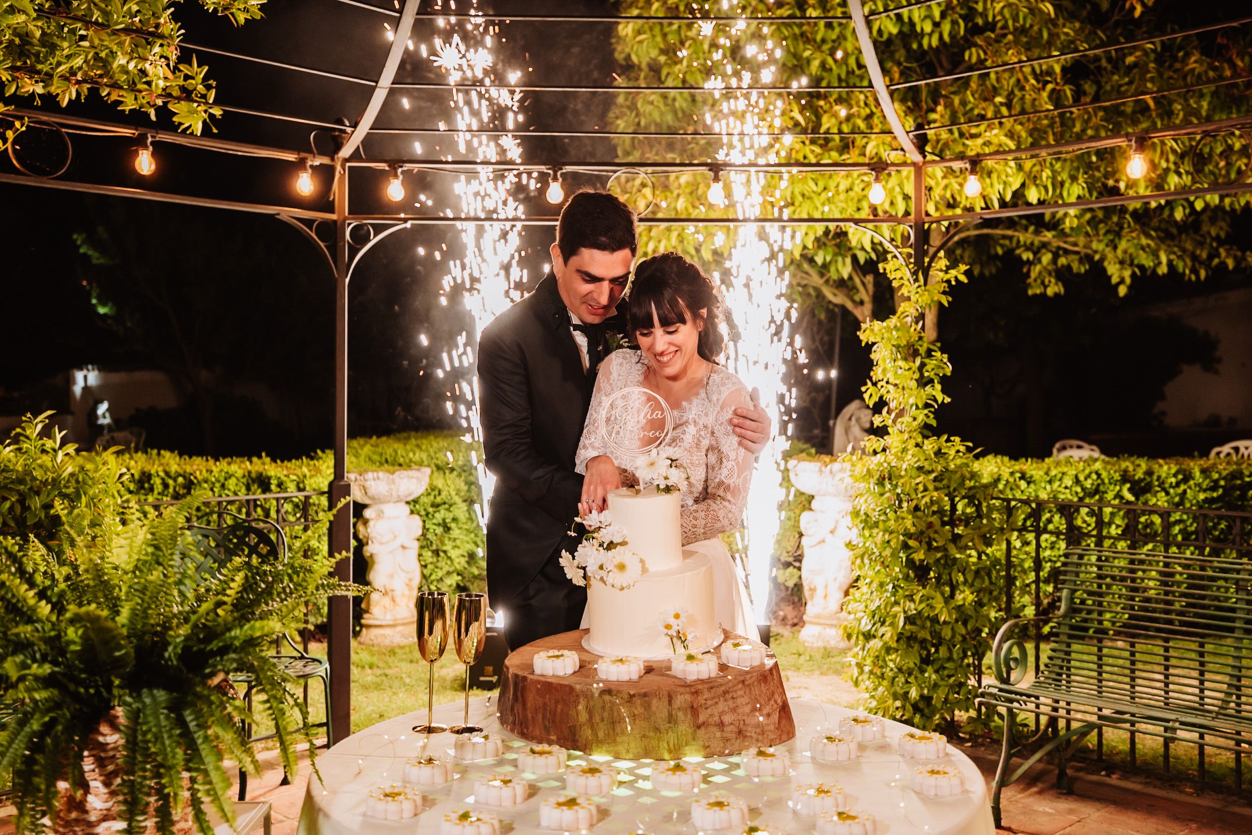 Bride and groom cutting wedding cake outdoors at night with sparkling fireworks and string lights in garden.