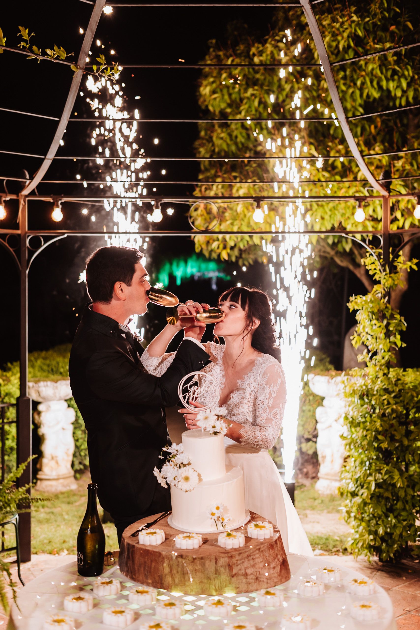 Bride and groom toasting champagne by their white wedding cake under sparkling fireworks and string lights at night.