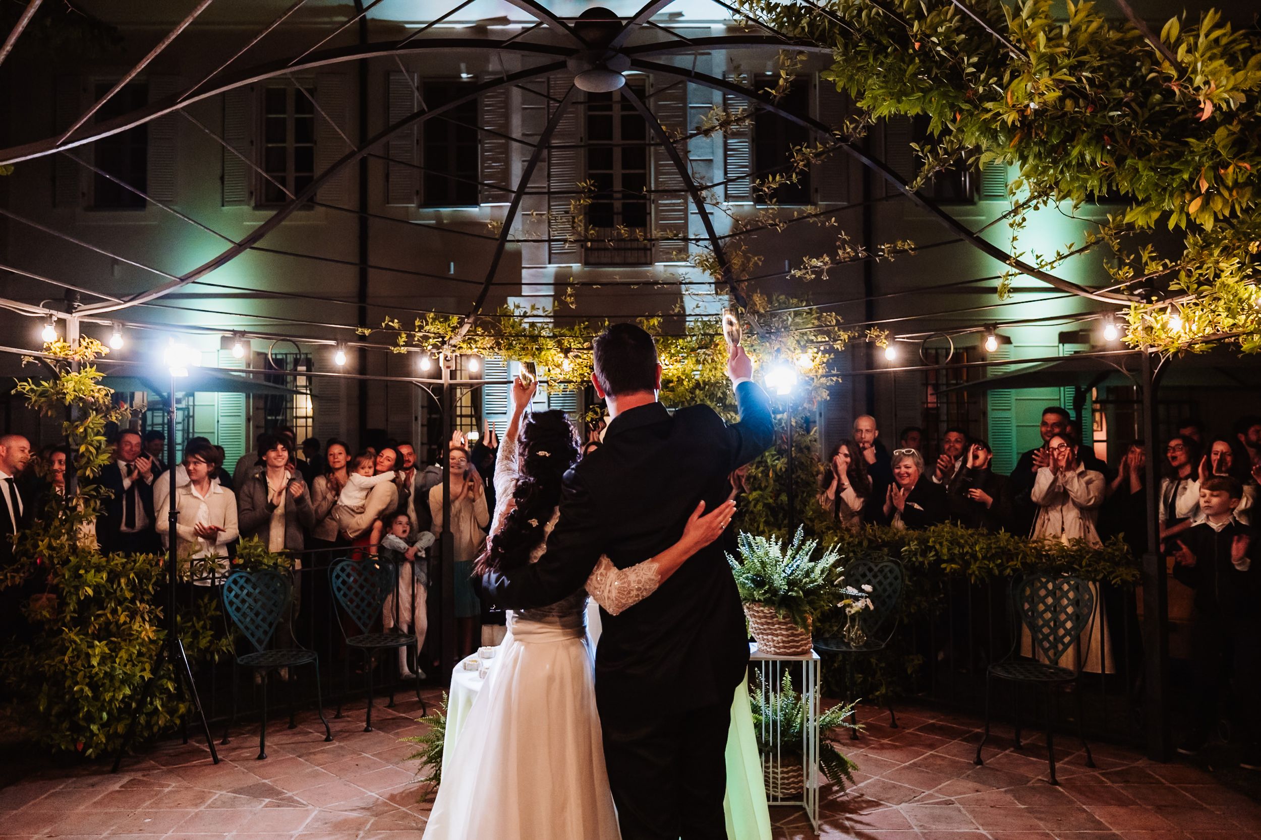 Bride and groom share first dance in romantic outdoor courtyard with string lights and applauding wedding guests at night.