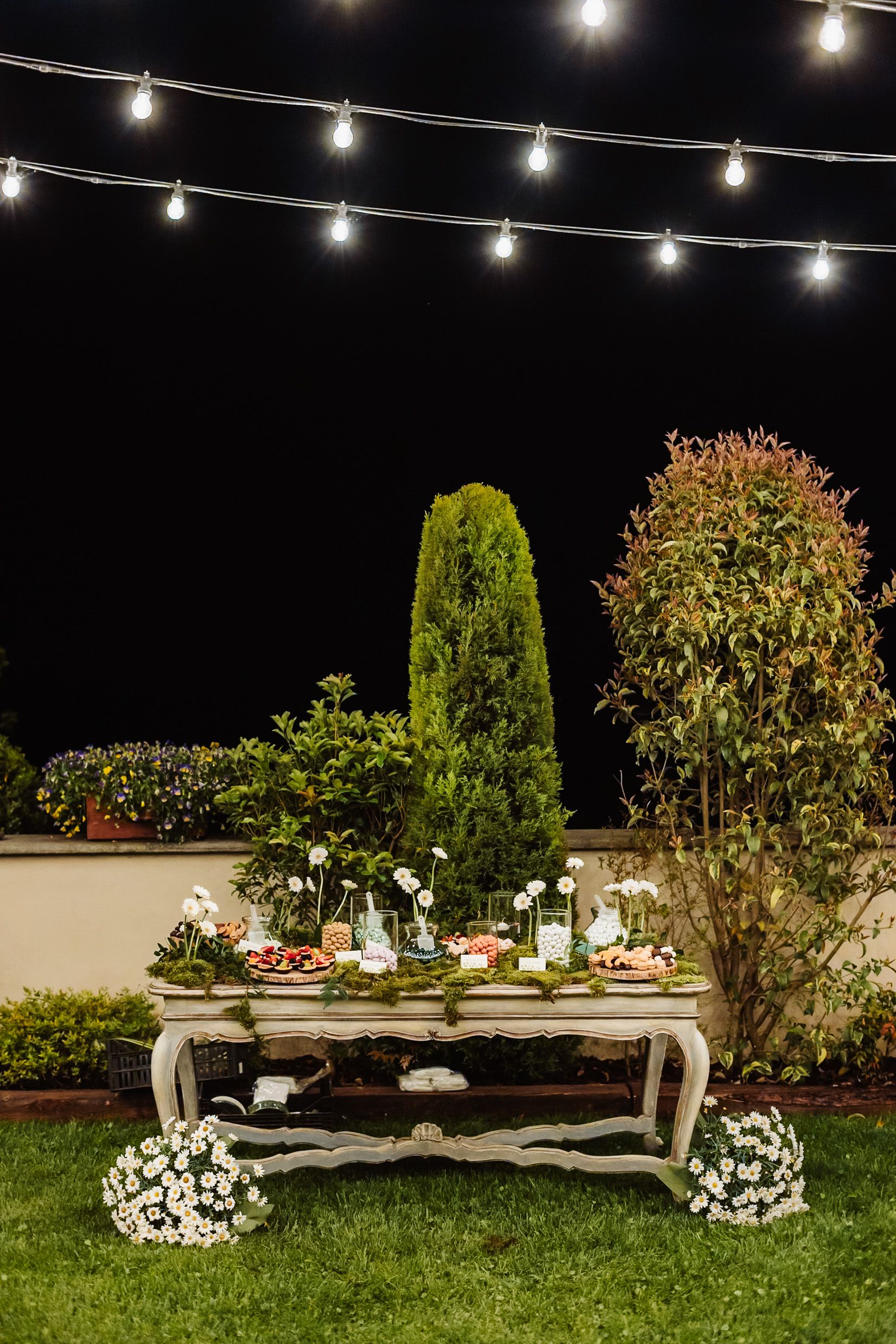 Elegant outdoor dessert table decorated with flowers and string lights in a garden at night.