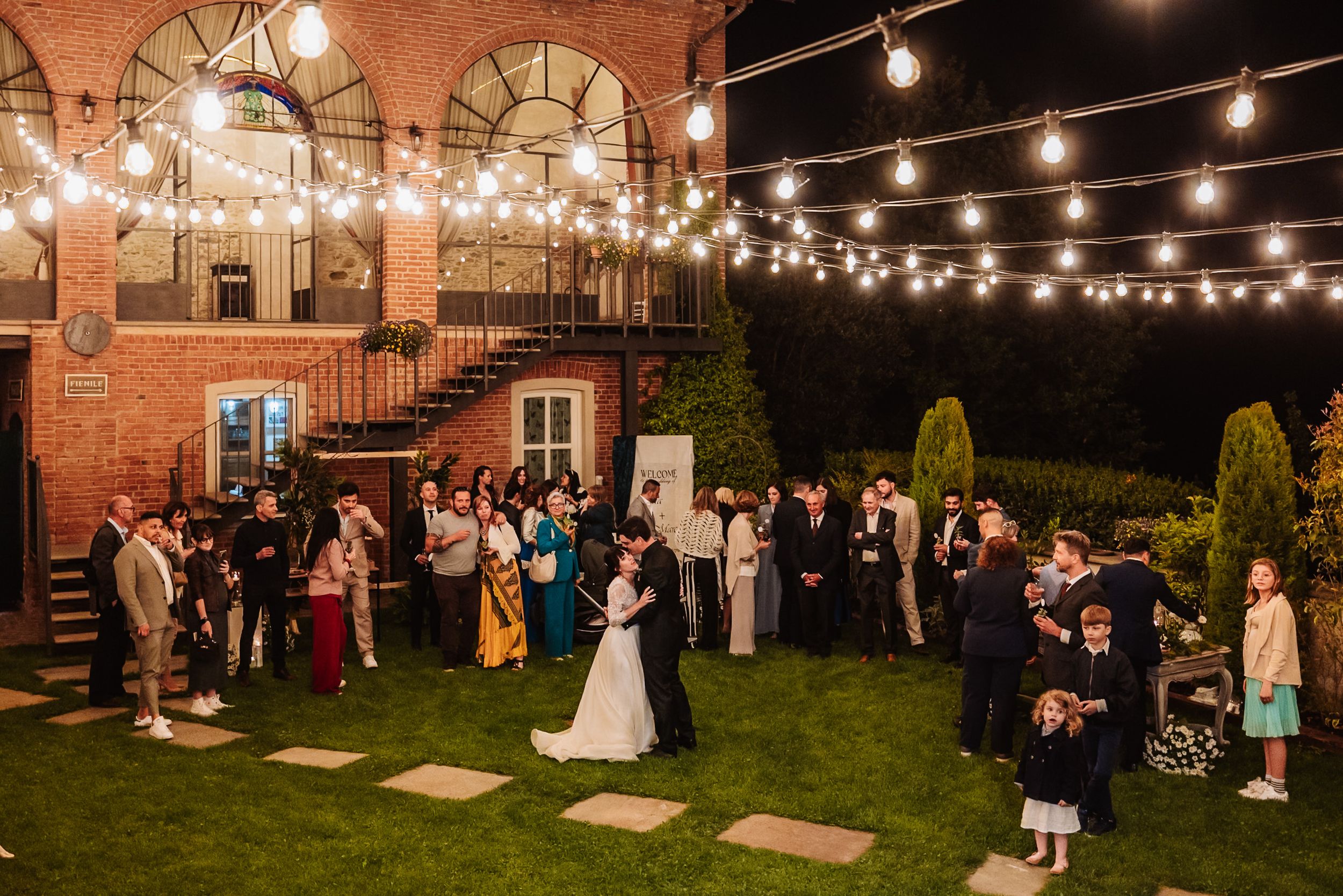 Outdoor wedding reception at night with string lights, bride and groom dancing surrounded by guests on a brick venue patio.