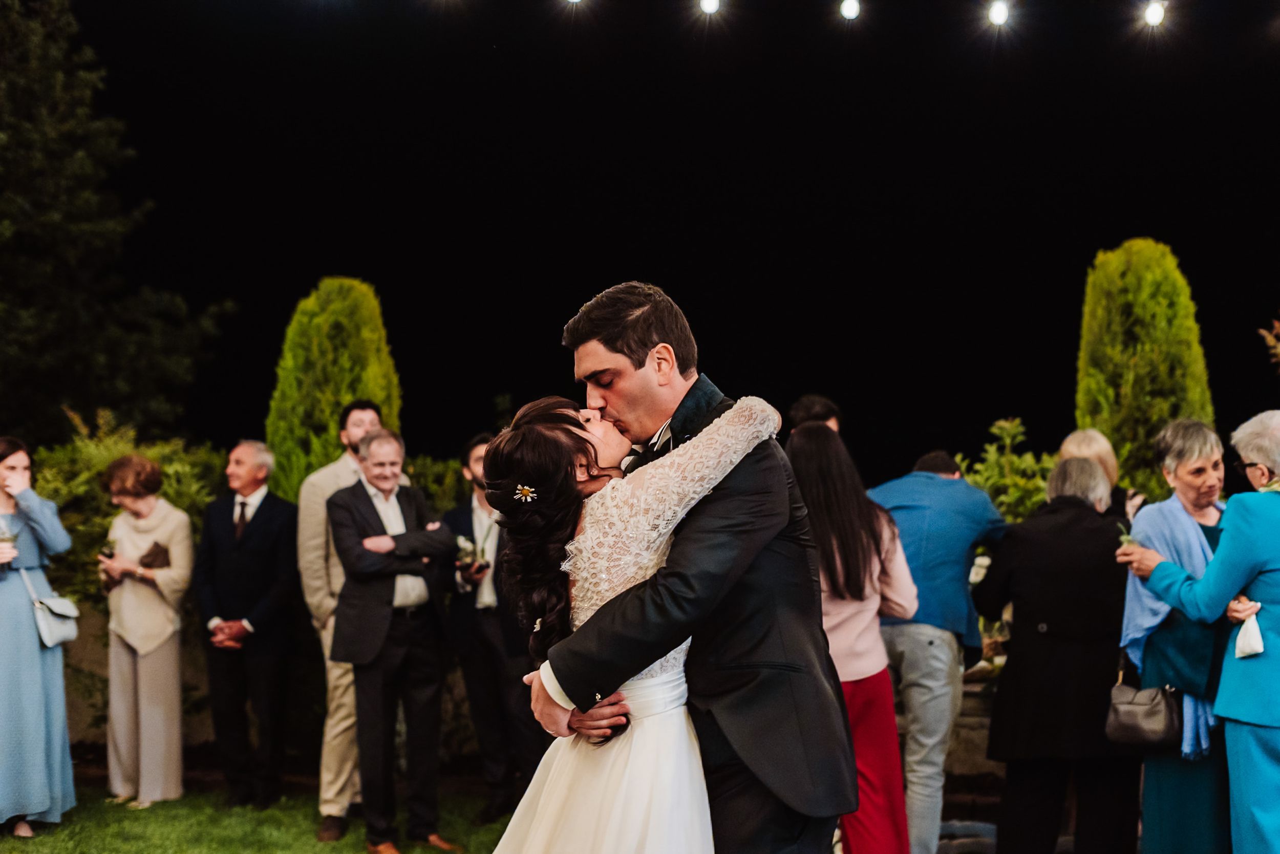 Bride and groom share first dance embrace at outdoor evening wedding reception with guests gathered around.