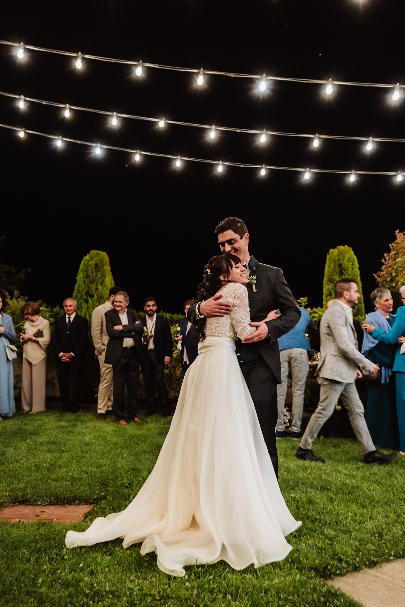 Bride and groom share first dance outdoors at night under string lights with wedding guests watching.