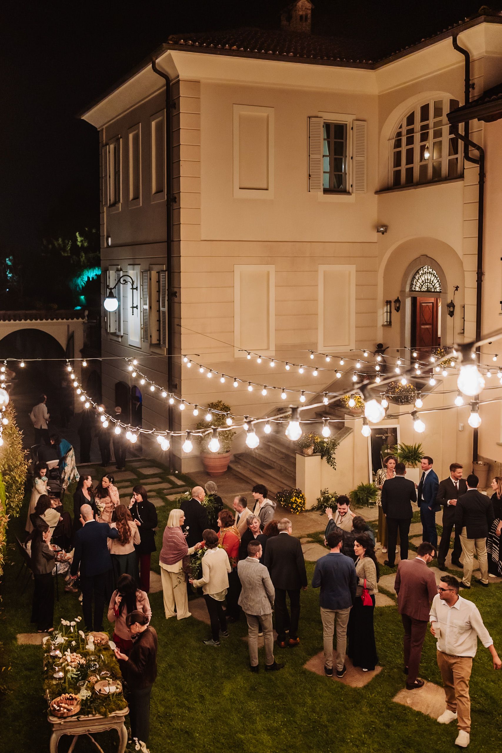 Elegant outdoor evening party with string lights at a villa, guests mingling on a lush lawn at night.