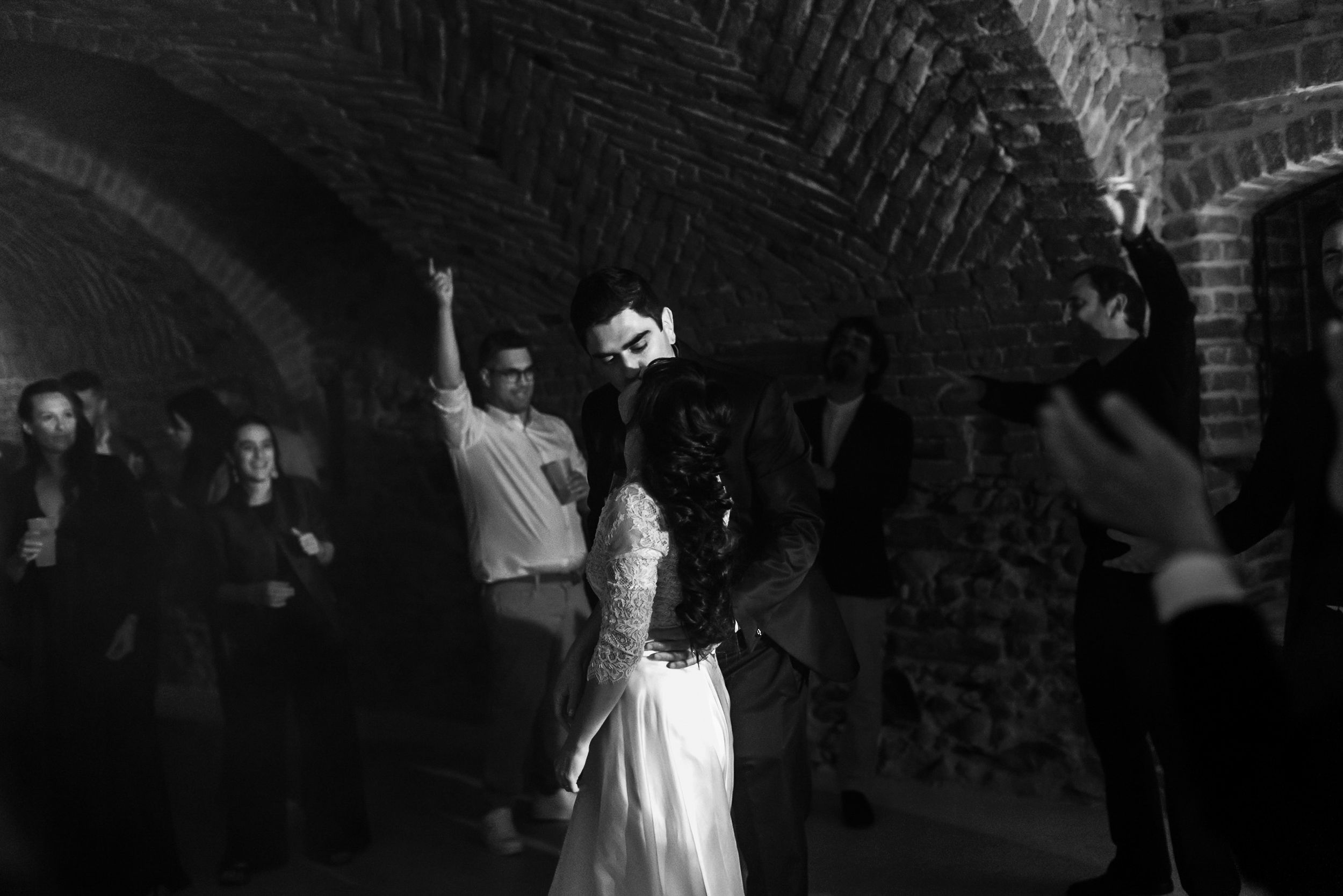 Black and white photo of a bride dancing with guests in a brick vaulted venue during a wedding reception.