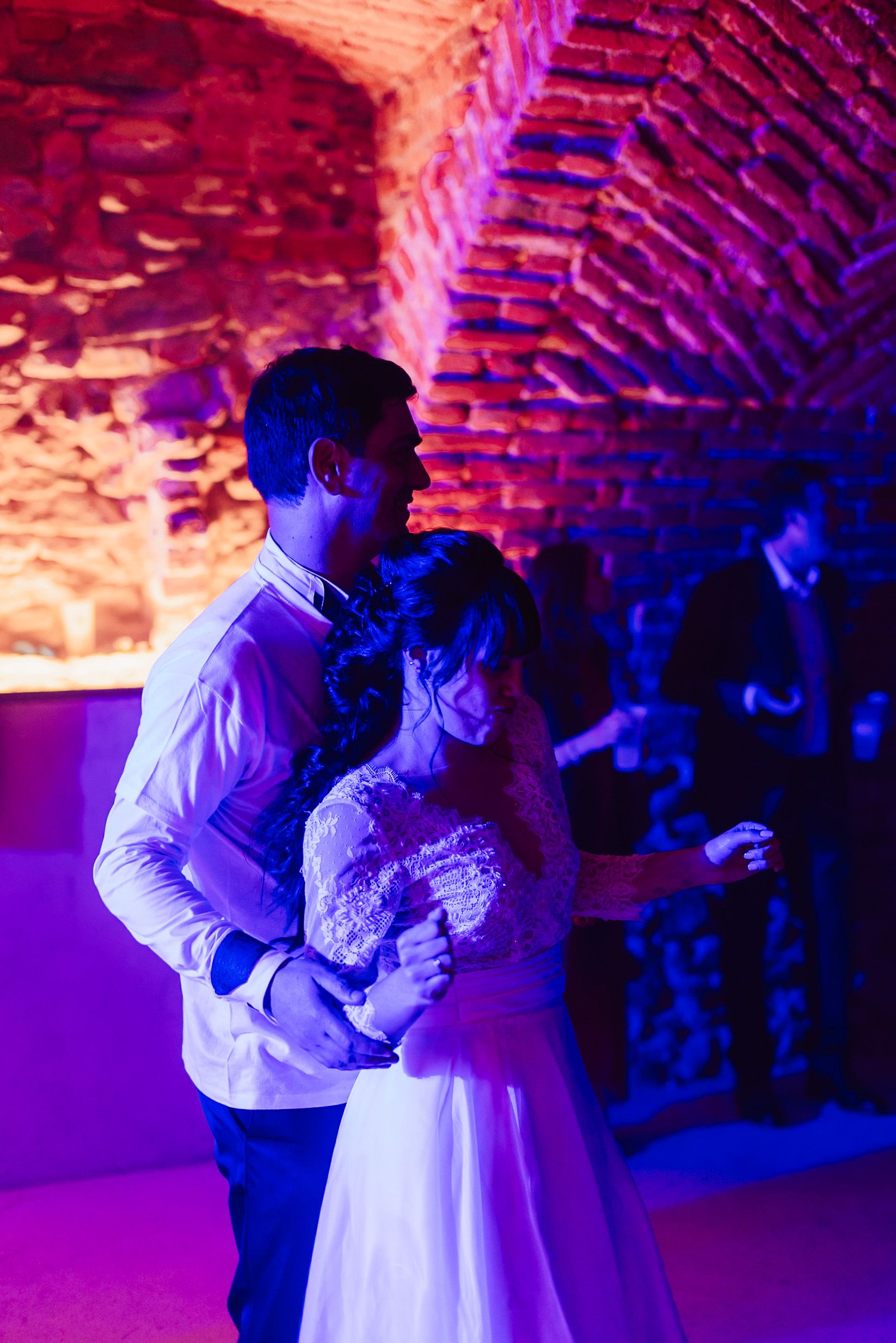 Newlyweds share romantic first dance in purple-lit stone venue during wedding reception.