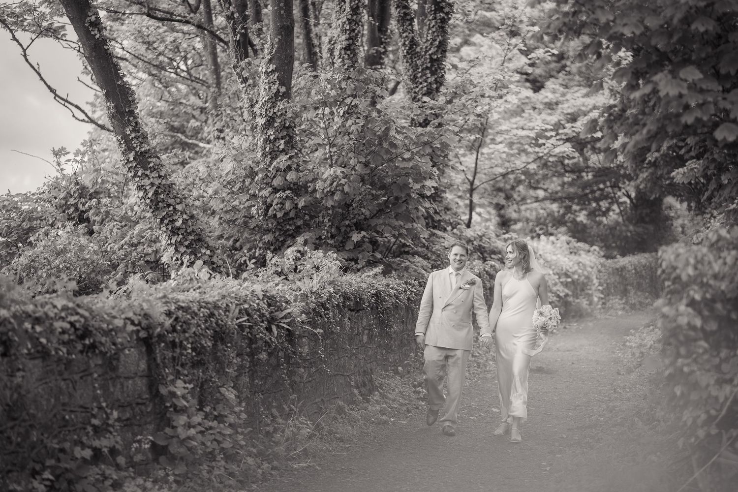 Black and white photo of a couple holding hands walking along a tree-lined path on their wedding day.
