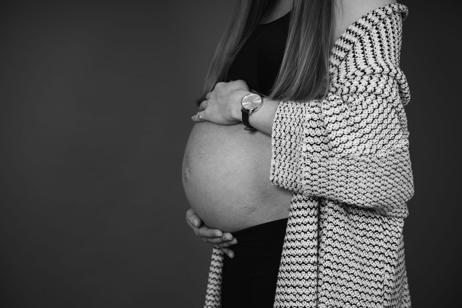 Black and white photo of a pregnant woman cradling her baby bump, wearing a knitted cardigan.