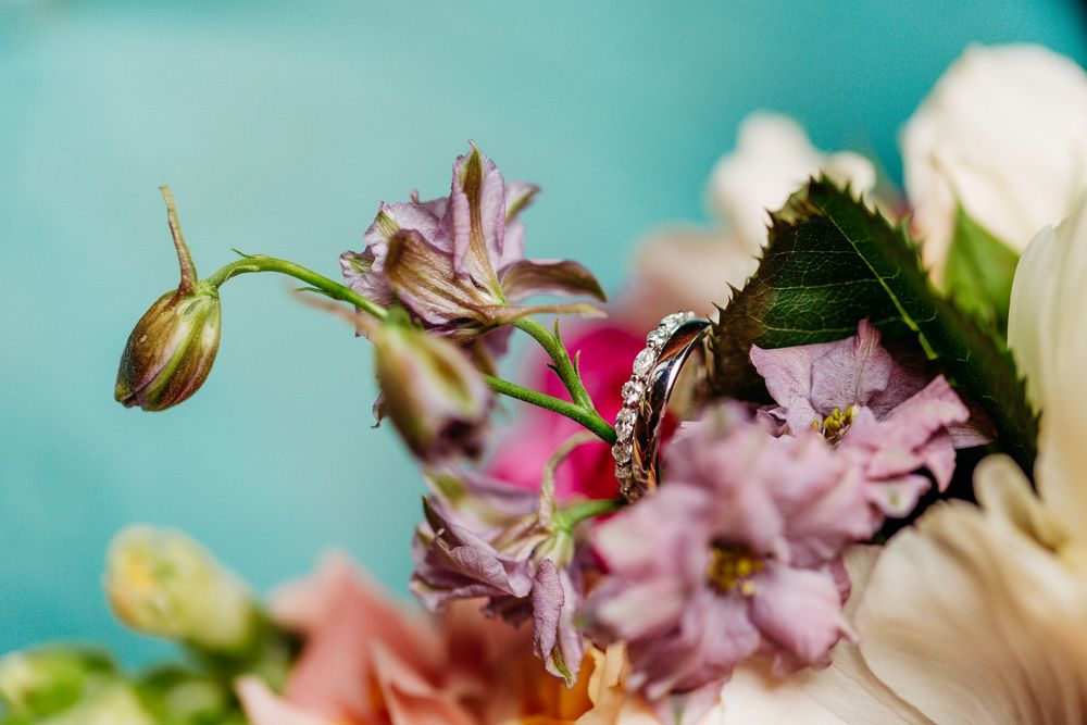 Diamond wedding ring resting on colorful spring flowers with pink and purple blooms against teal background.