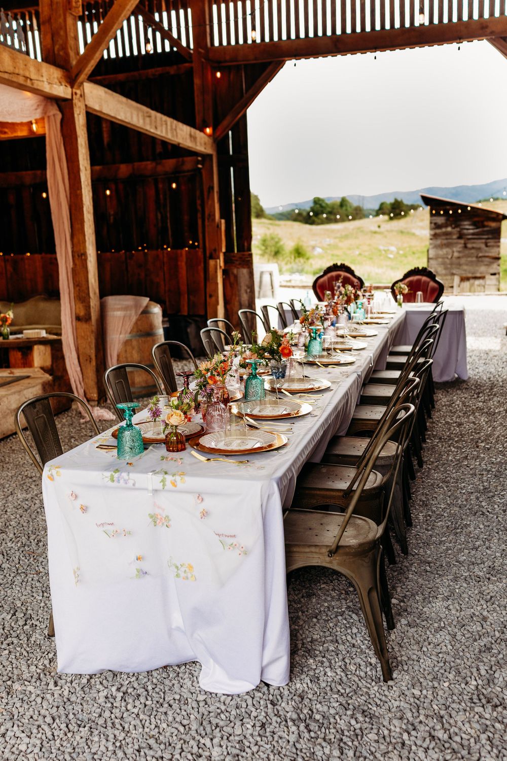 Elegant rustic barn wedding reception with long table, floral centerpieces, mixed metal chairs, and string lights.