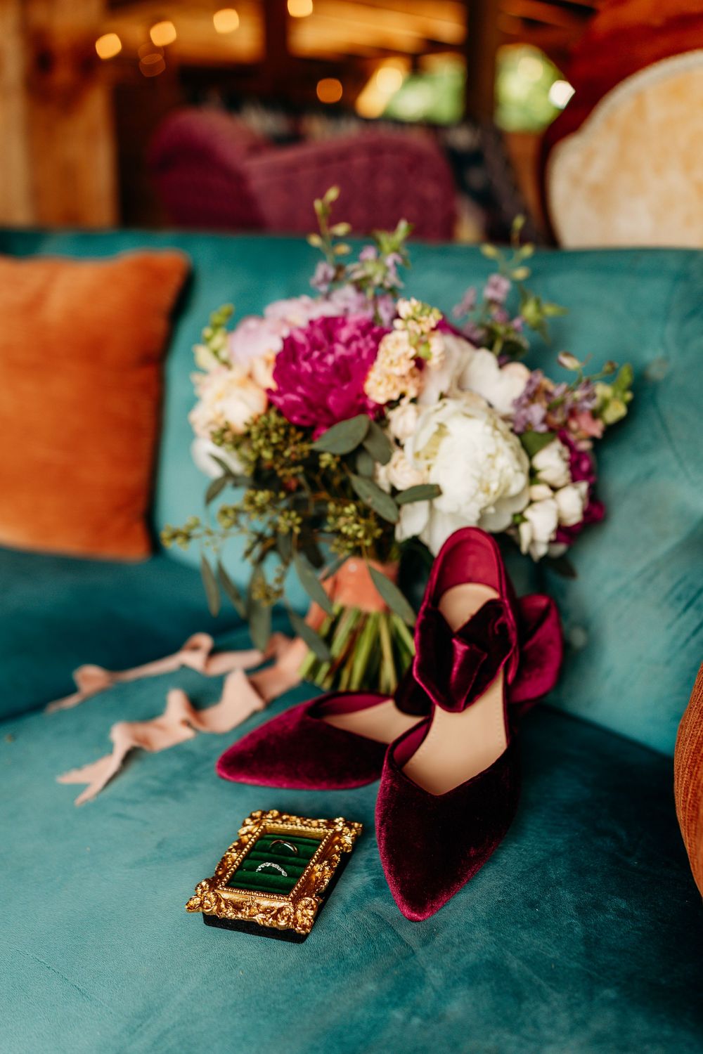 Burgundy velvet heels, floral bouquet, and gold ring box on teal velvet sofa for elegant wedding styling.