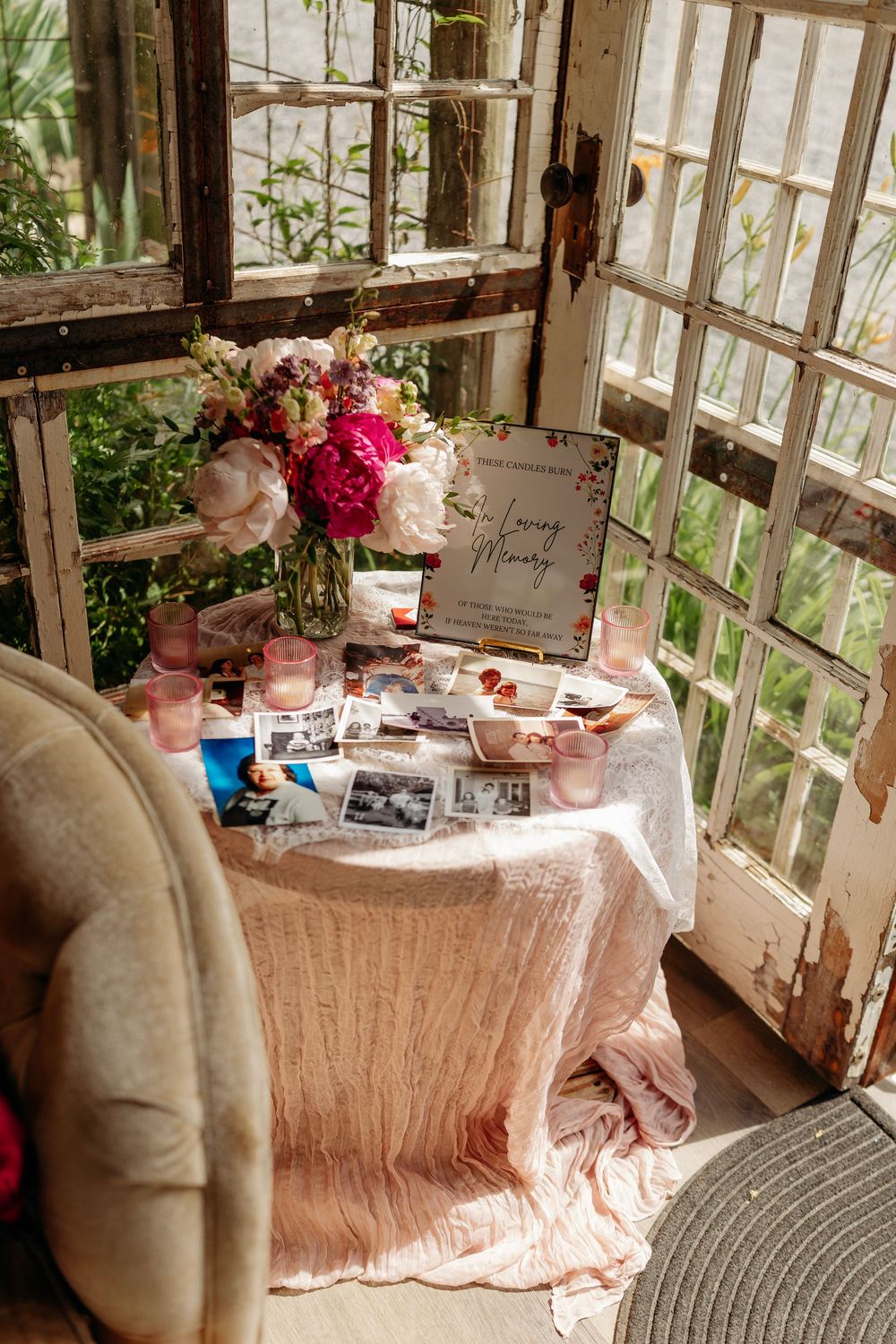 A memorial table with pink flowers, candles, and black-and-white photos in a rustic greenhouse setting.