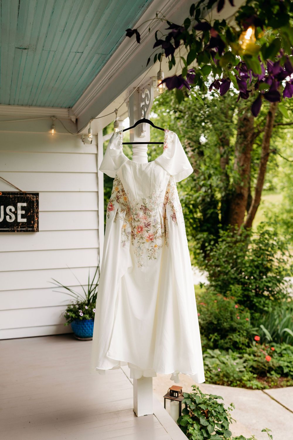 White wedding dress with floral embroidery hanging on a porch hanger surrounded by lush greenery and purple flowers.