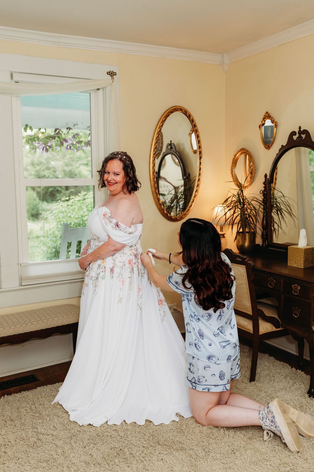 Bride in floral wedding gown being helped by bridesmaid in vintage bedroom with gold mirrors.