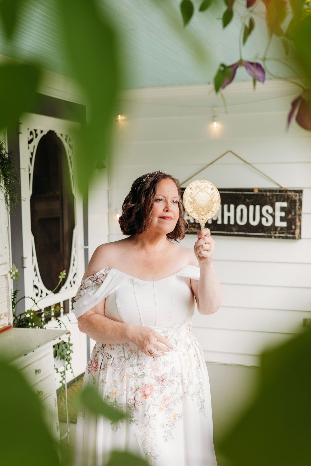 A bride in an off-shoulder floral gown holds a vintage mirror on a charming white porch with string lights.