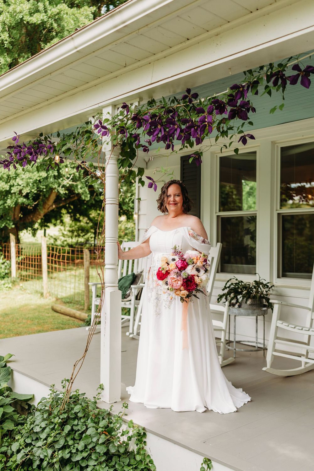 Bride in off-shoulder white gown holding vibrant bouquet on a charming farmhouse porch with purple clematis.