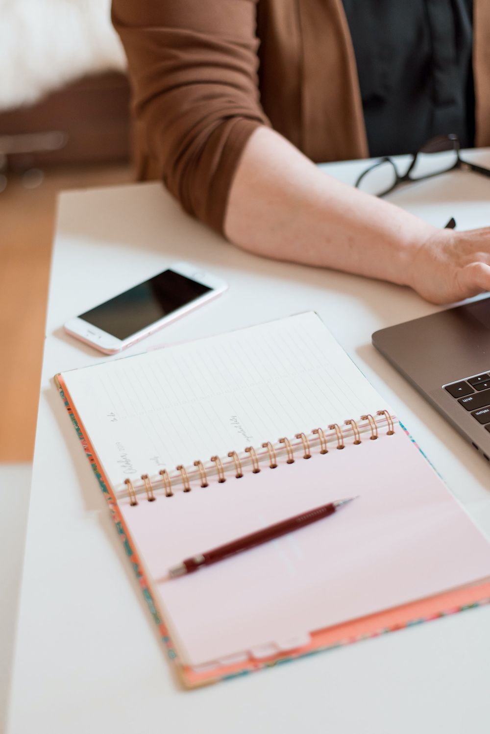 Workspace setup with spiral notebook, pencil, smartphone and laptop on white desk.