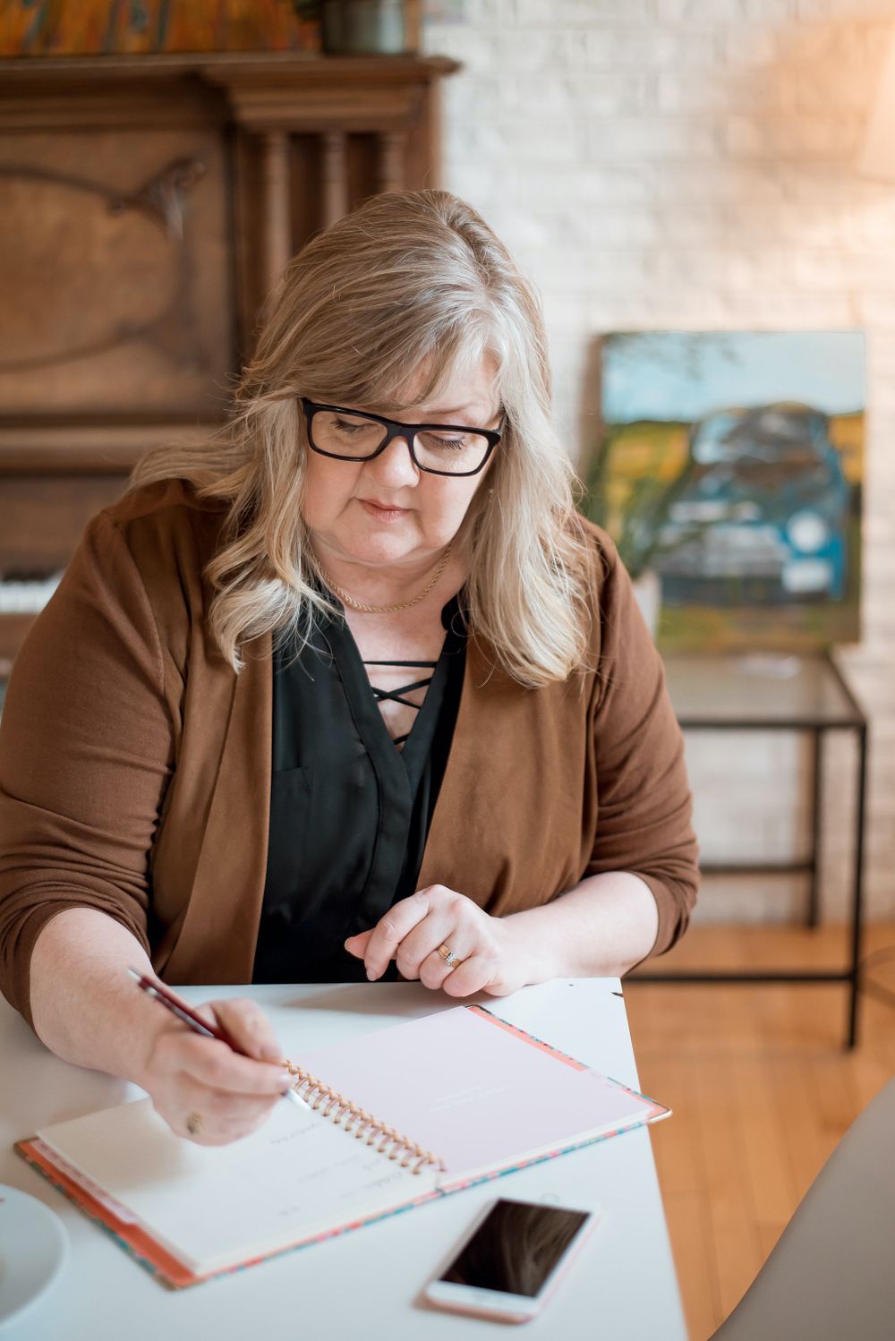 Person writing in notebook at desk with smartphone and artwork in background.