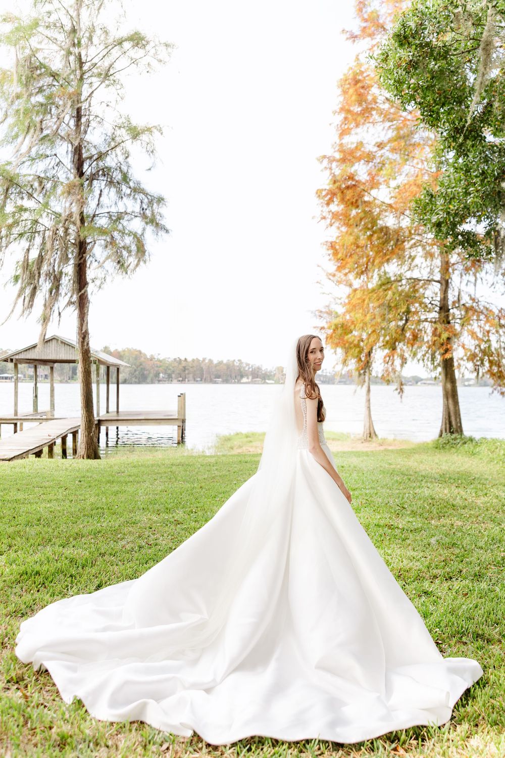 Bride in elegant white ball gown with long train stands on green lawn by lakeside dock surrounded by autumn trees.