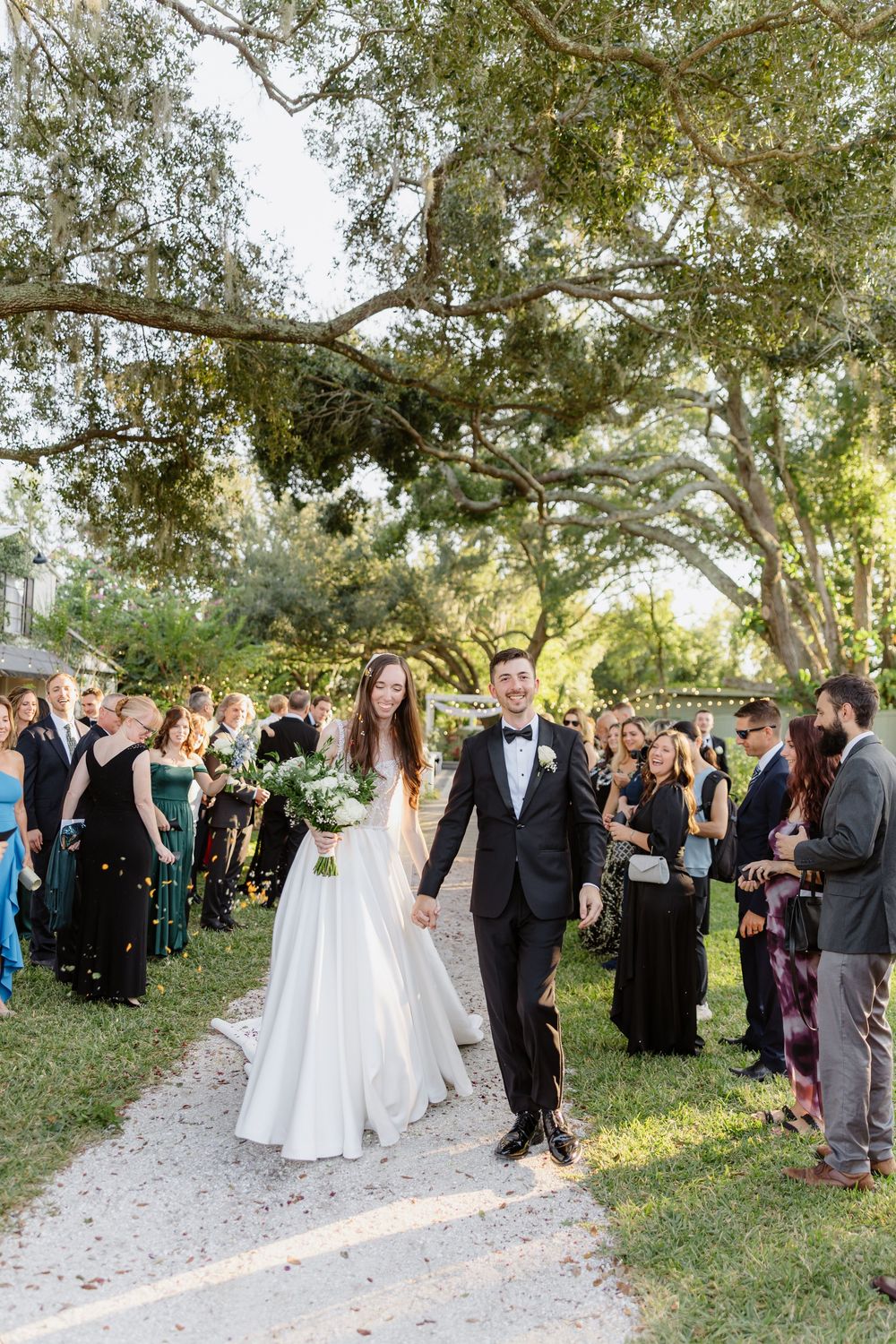 Bride and groom walk down outdoor wedding aisle holding hands while guests celebrate among lush green trees.
