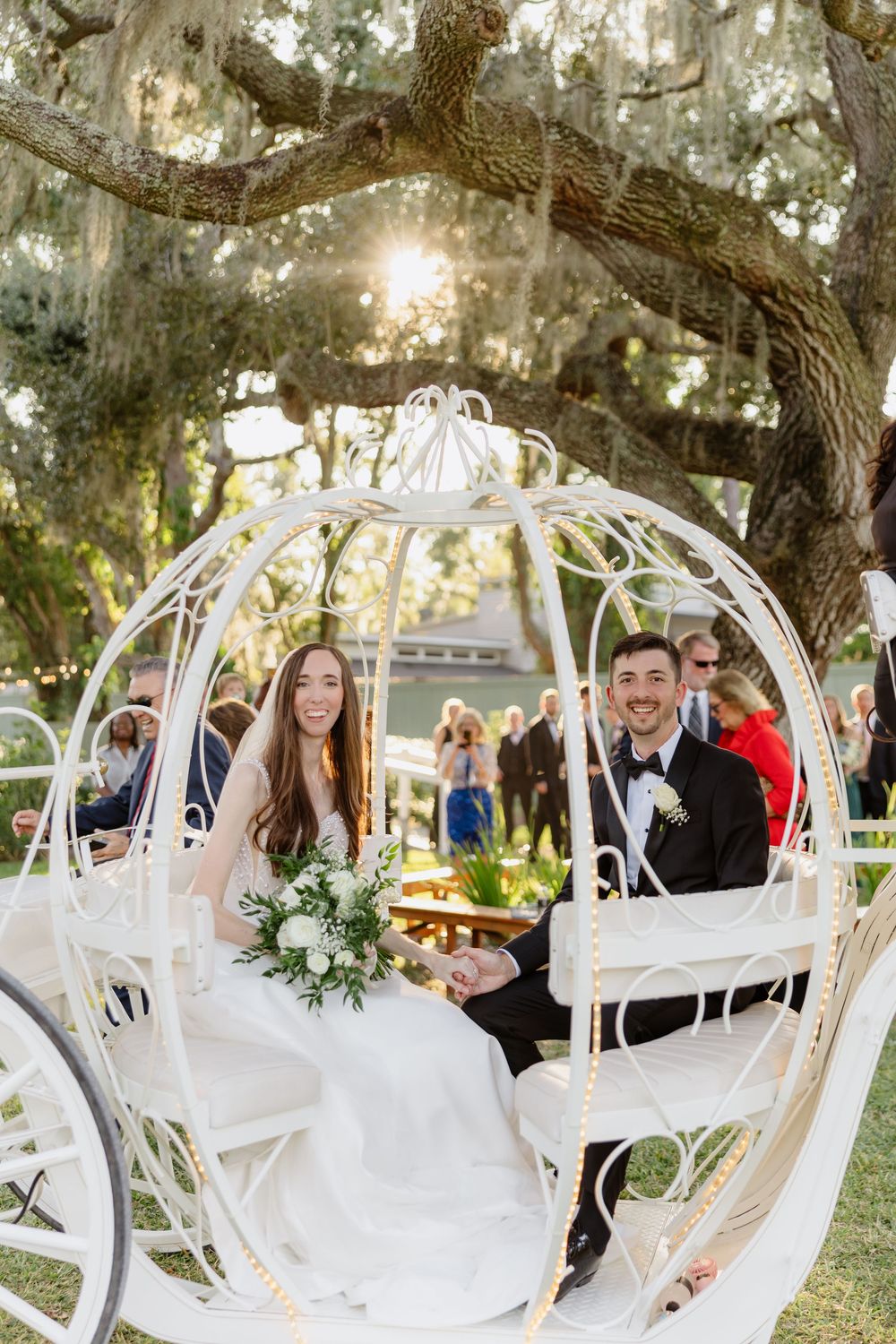 Bride and groom smile in a white Cinderella carriage under a large oak tree at an outdoor wedding.