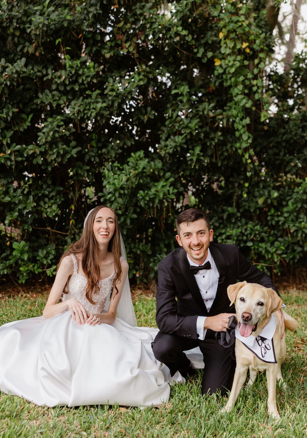 Smiling bride and groom in wedding attire pose outdoors with their yellow Labrador wearing a bow tie bandana.
