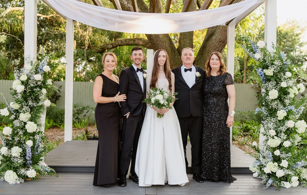 Wedding party of five poses under white pergola with floral arrangements at elegant outdoor ceremony.