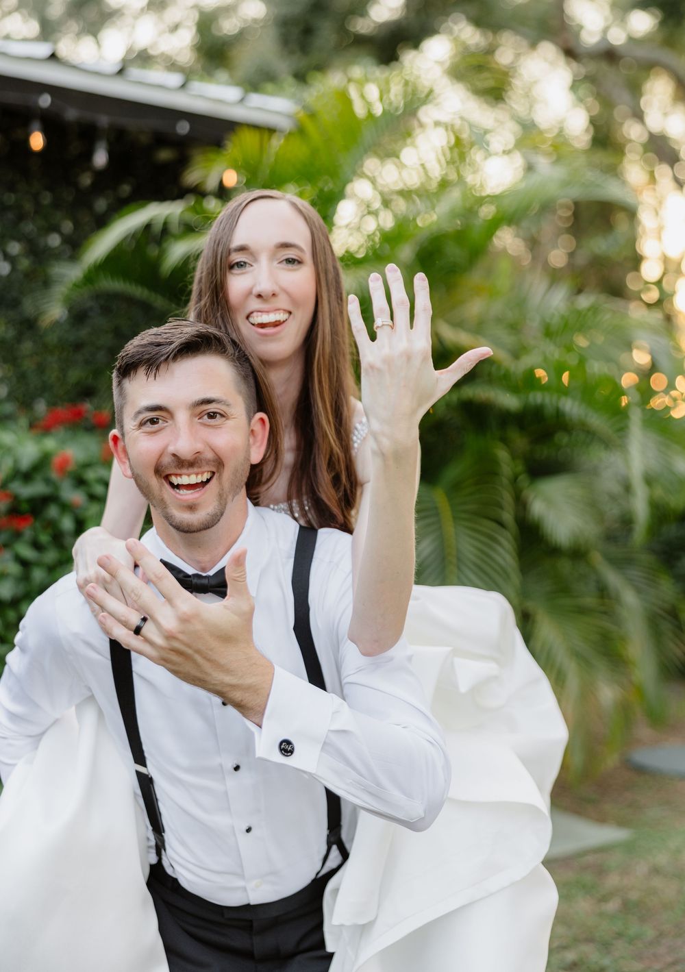 Laughing groom carries bride on his back outdoors as she shows off wedding ring to camera.