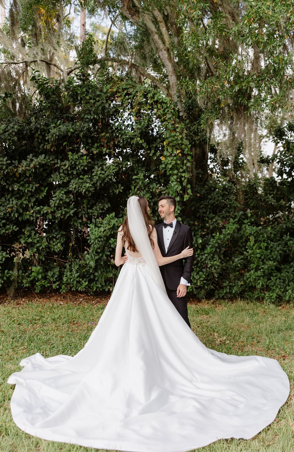 Bride in elegant white gown with long train stands with groom in black tuxedo under lush green trees outdoors.