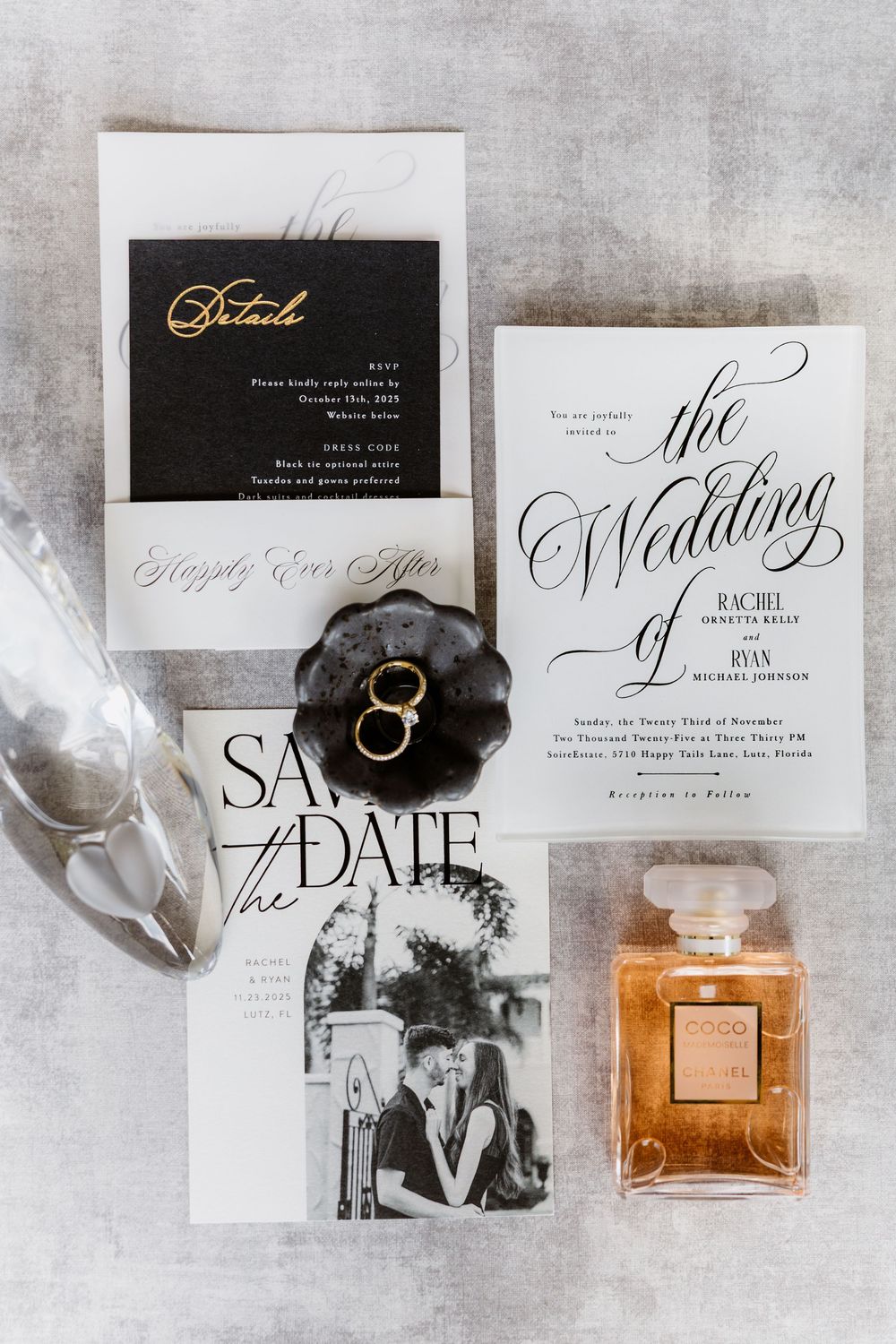 Elegant black and white wedding invitation suite with gold details, rings, and Chanel perfume on gray surface.
