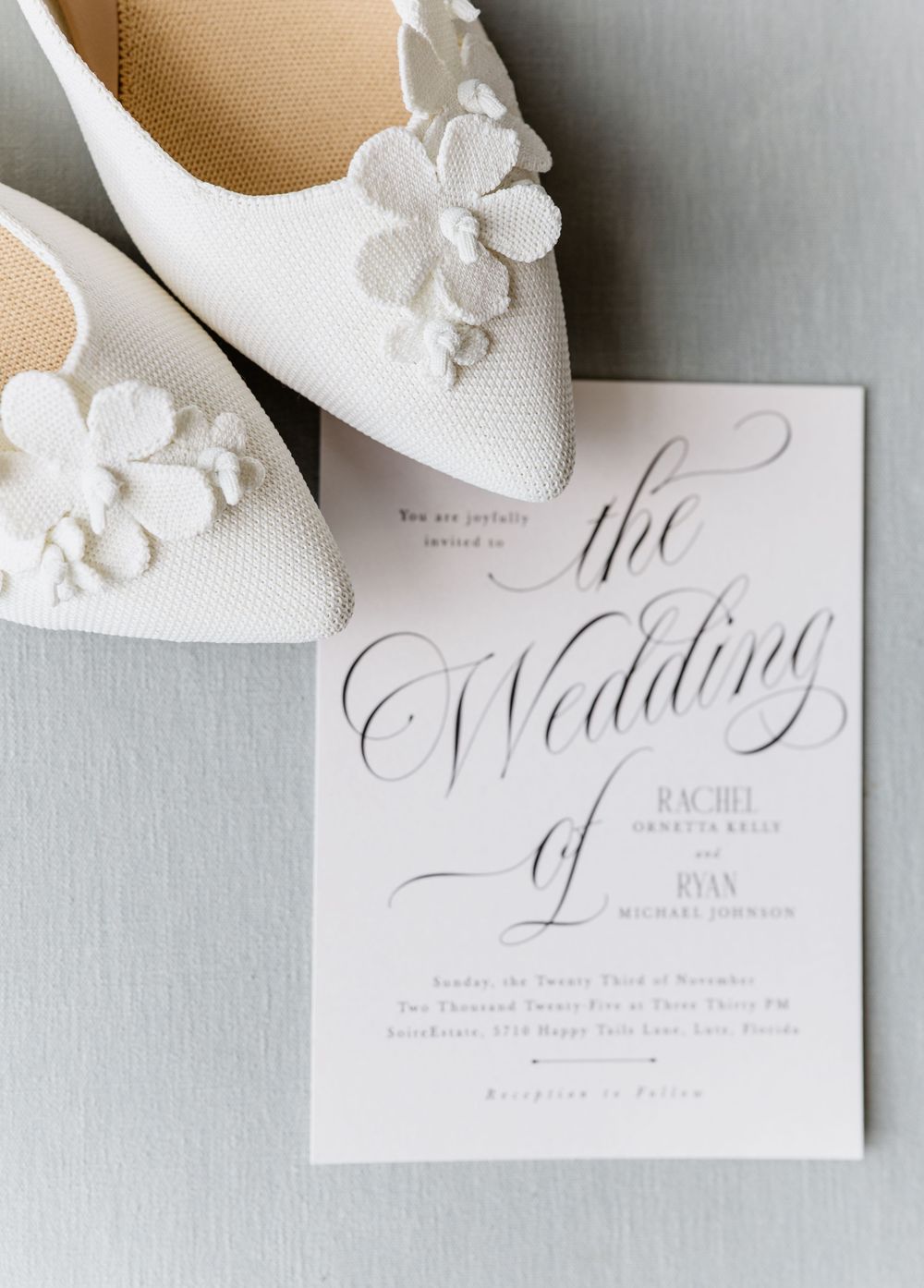 Wedding invitation for Rachel and Ryan beside white floral heels on a soft gray background.