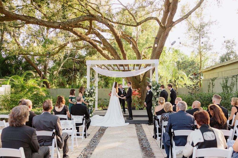Outdoor wedding ceremony under a sprawling oak tree with white arbor, aisle, and seated guests in attendance.