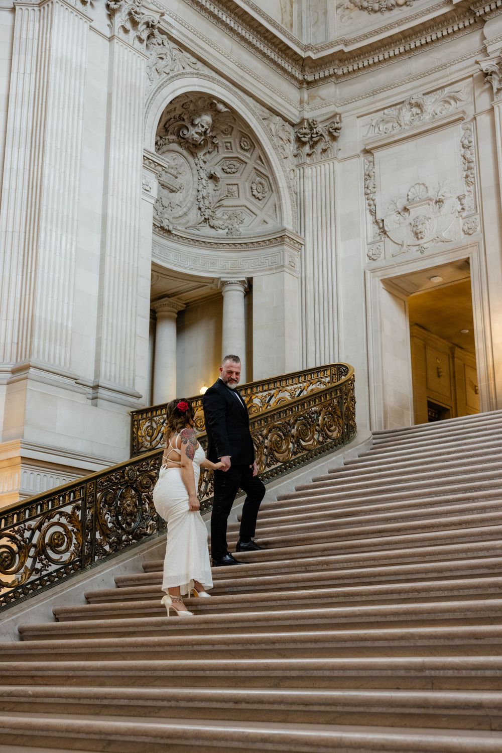 Couple in wedding attire descending grand marble staircase with ornate gold railing in elegant neoclassical building.