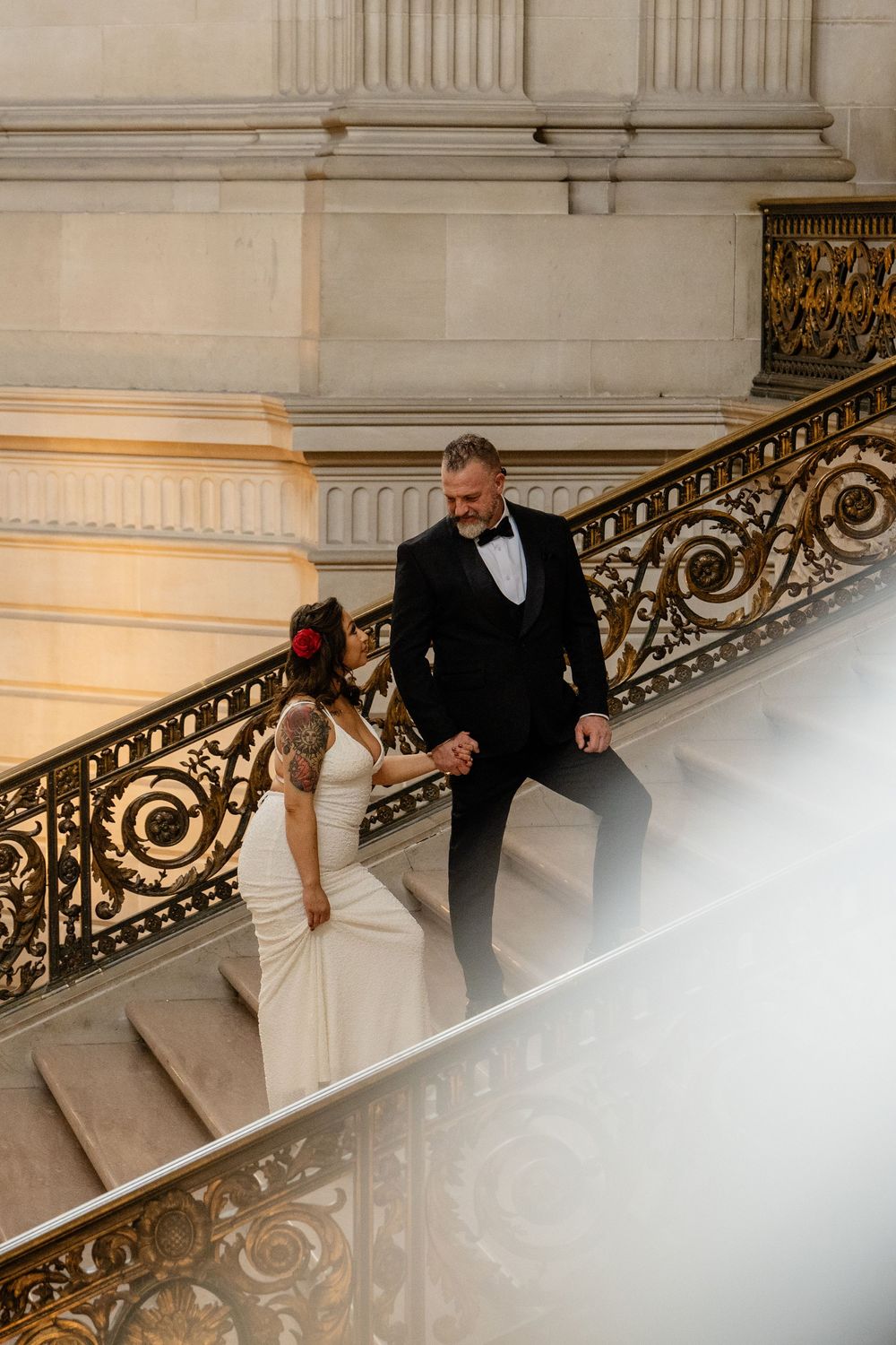 Bride and groom holding hands on ornate staircase, bride in white gown with red hair flower, groom in black tuxedo.
