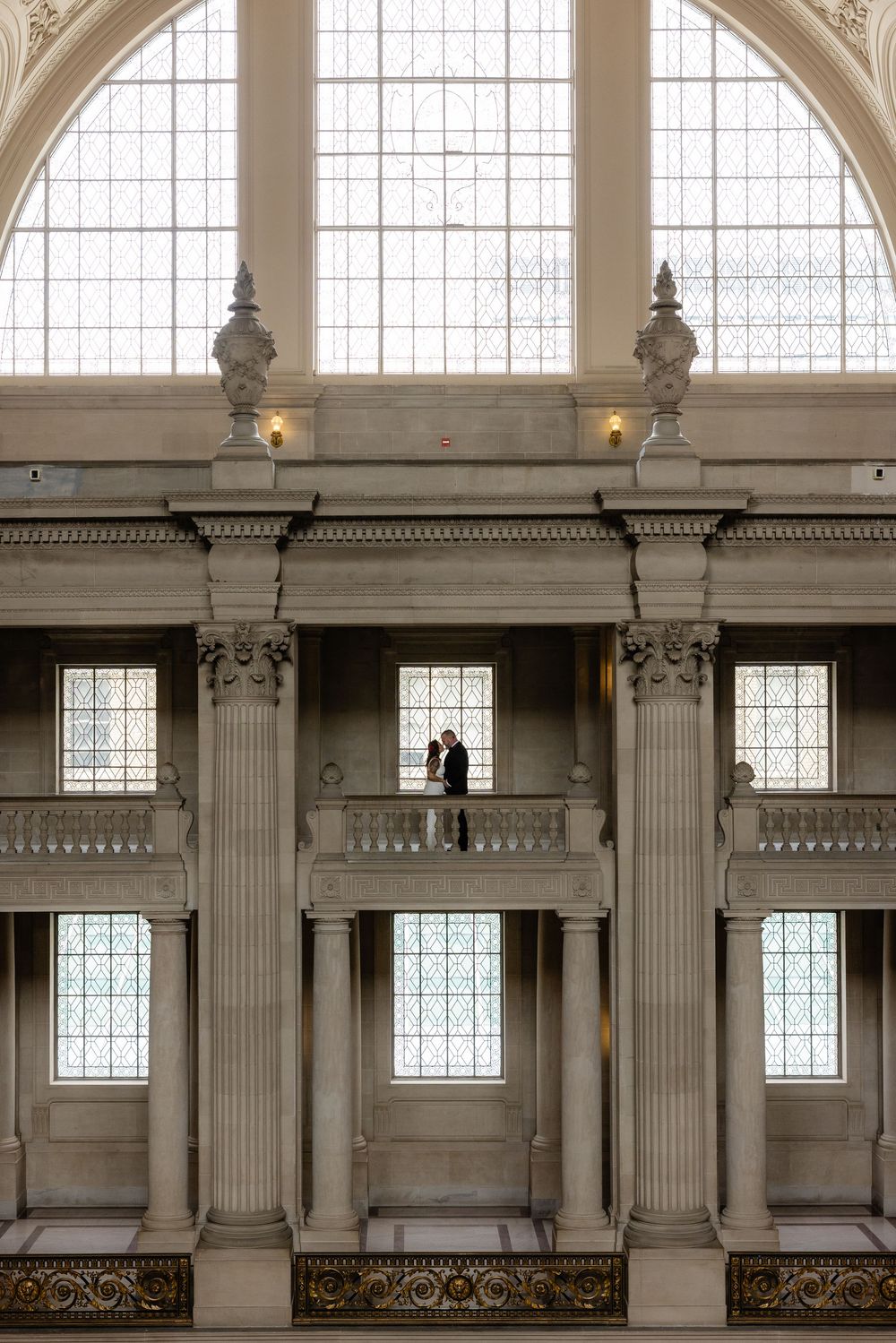 Couple silhouetted on a grand balcony inside San Francisco City Hall with tall columns and arched windows.