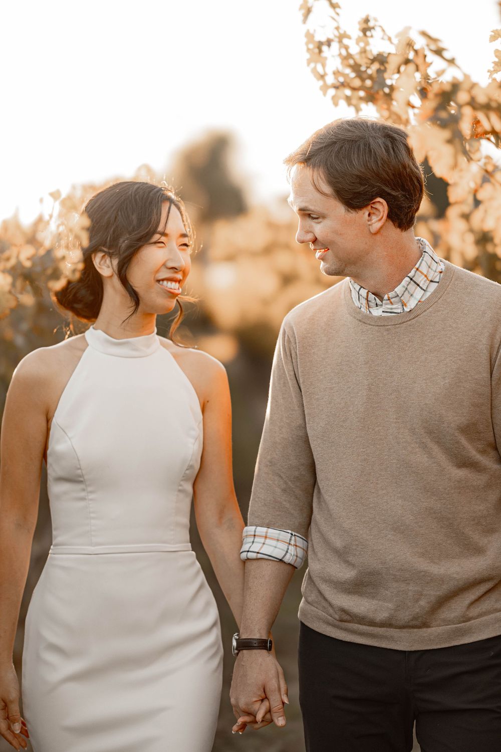 Happy bride and groom walk hand in hand through sun-drenched vineyard, gazing lovingly at each other.