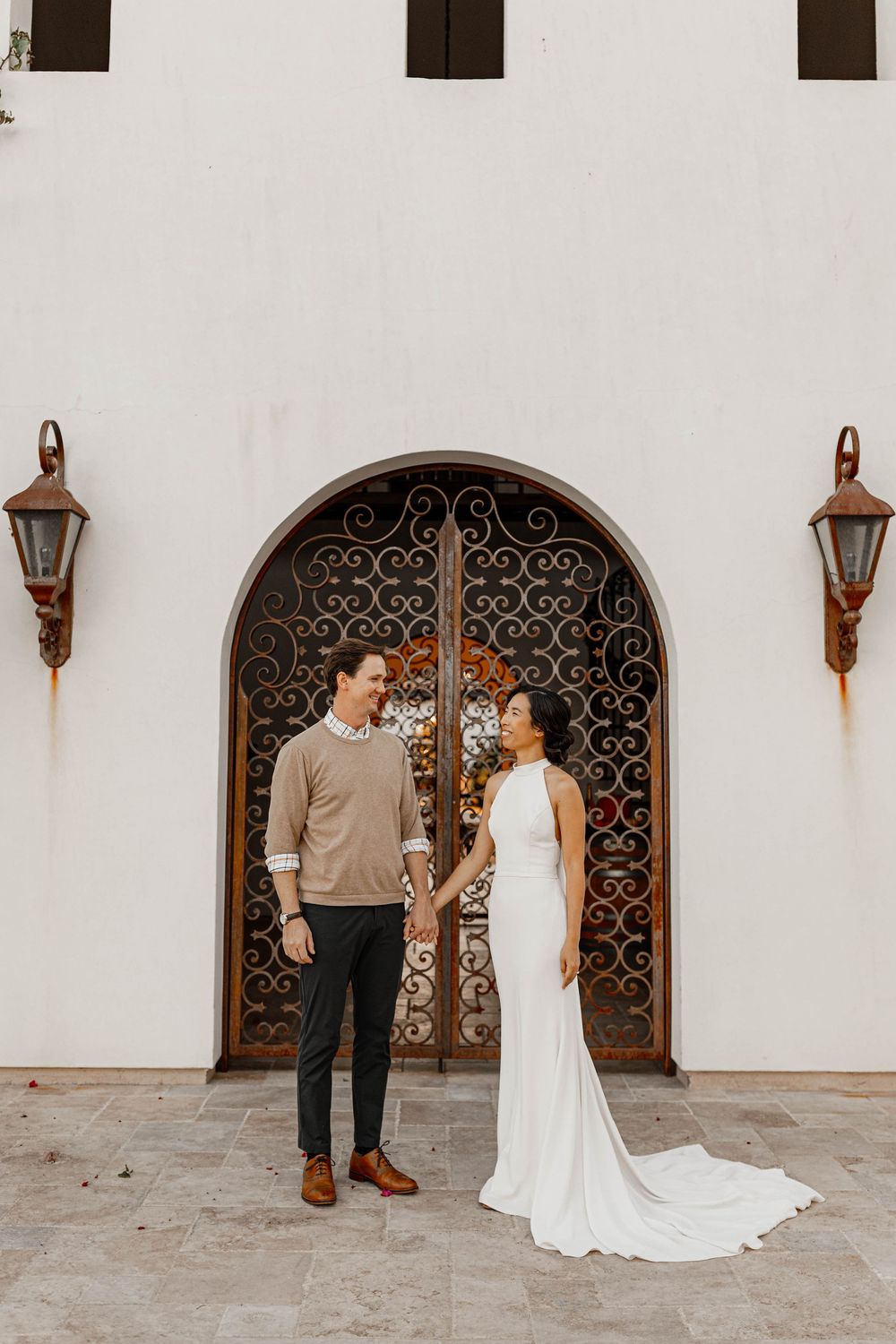 Couple holding hands before ornate iron doors of white Spanish-style building, bride in elegant gown.