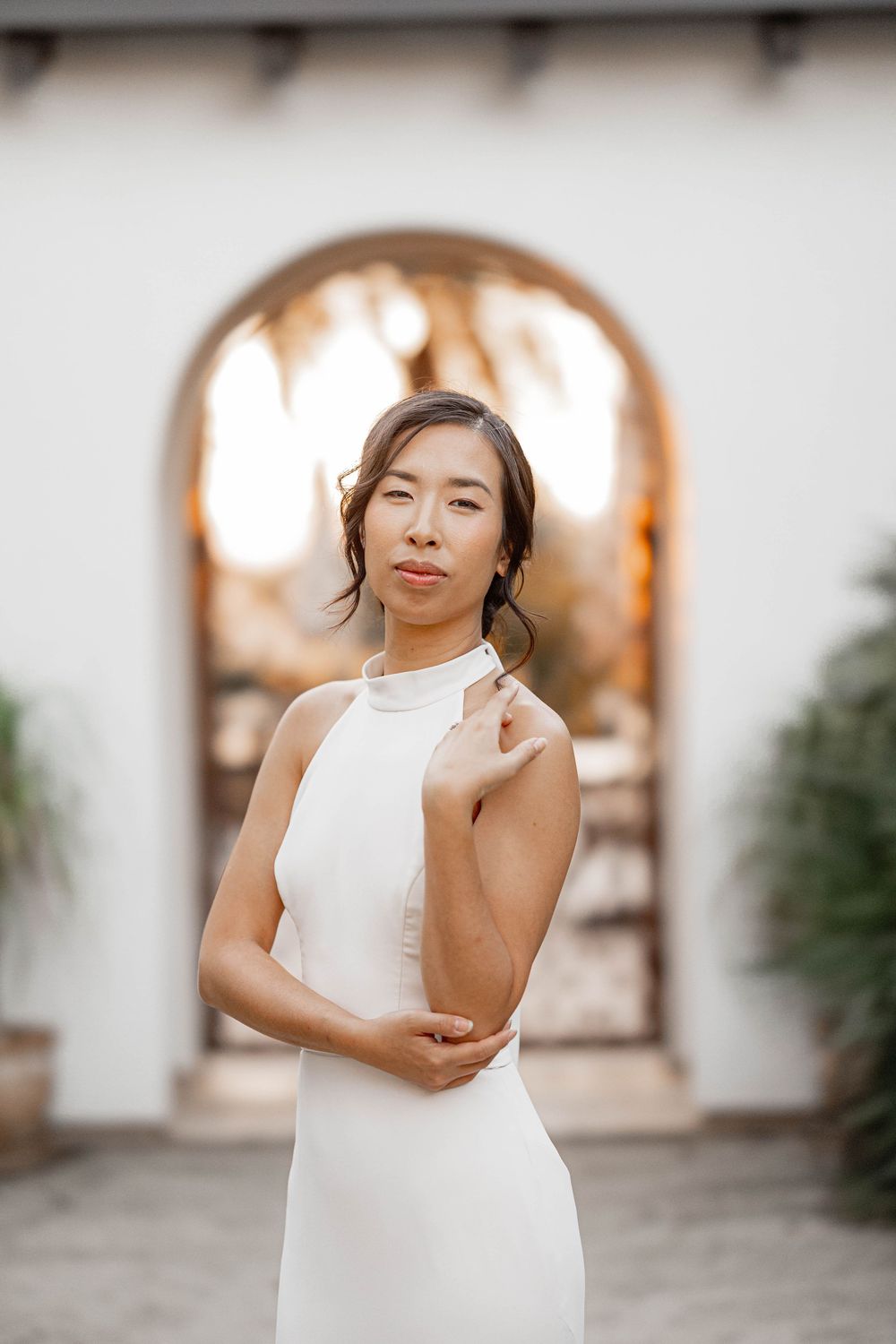 Elegant bride in white halter gown poses before arched doorway with warm golden light at outdoor venue.