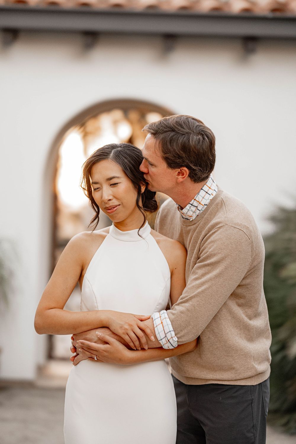 Groom kisses bride on cheek as she smiles in white halter gown near arched building.
