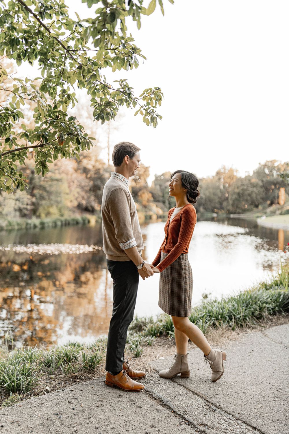 Couple holding hands by a serene autumn pond, surrounded by golden foliage in a romantic outdoor setting.