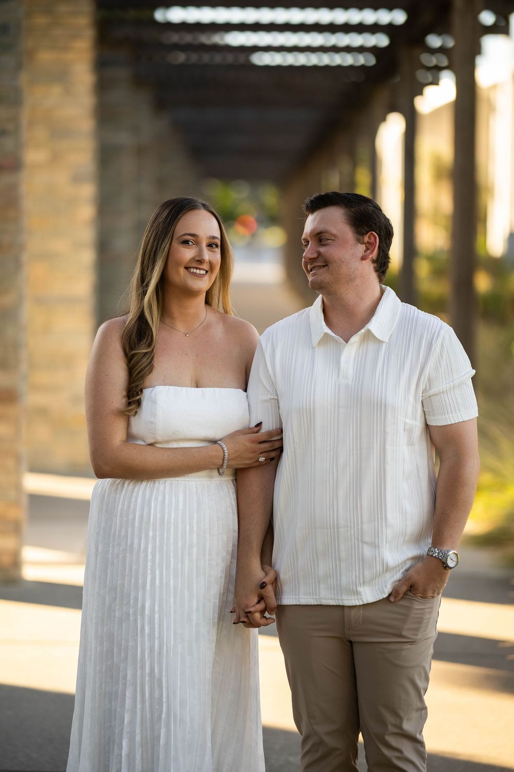 Smiling couple in matching white outfits holding hands under a pergola at golden hour.