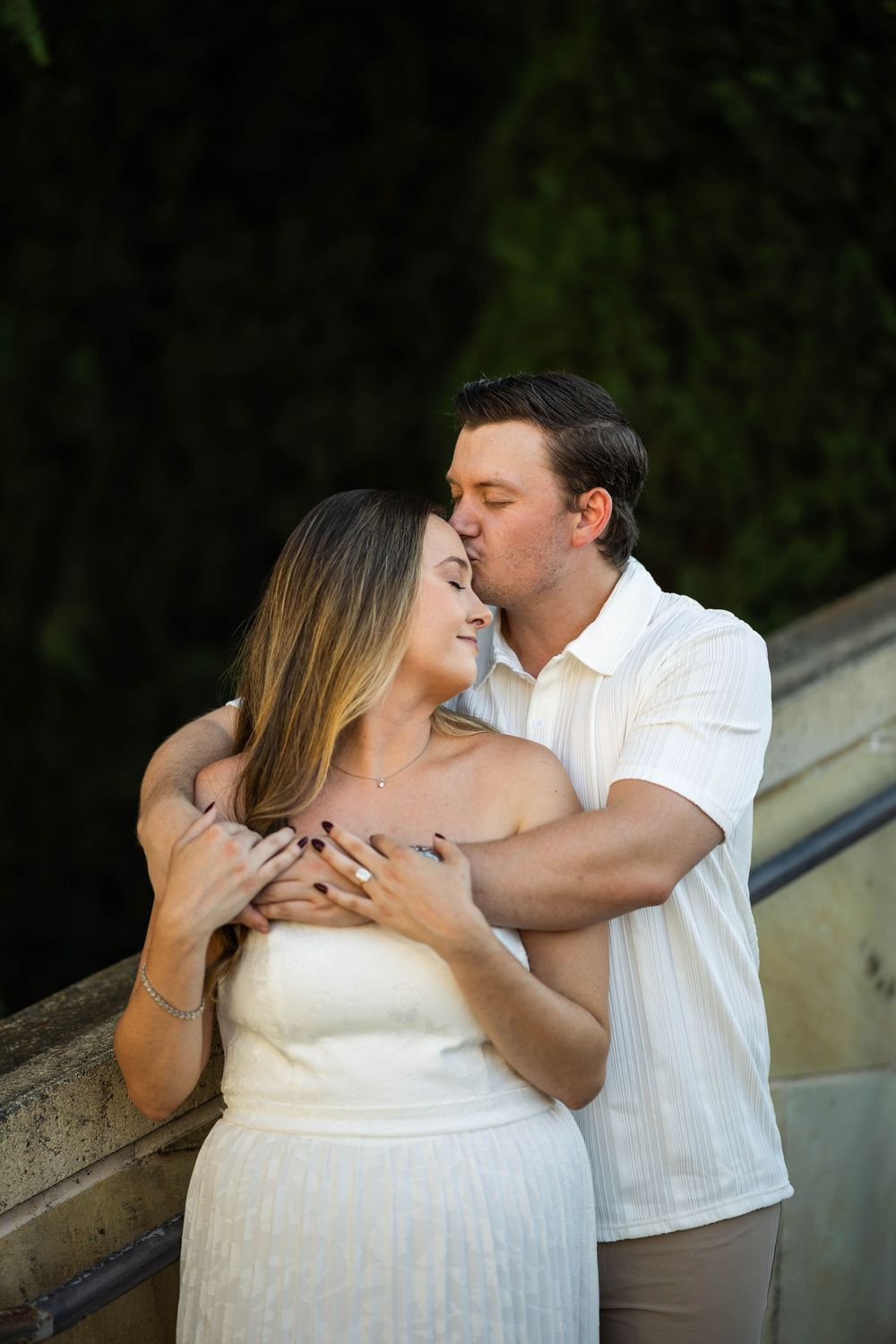 Couple in white outfits sharing a romantic kiss outdoors, man embracing woman from behind near stone railing.