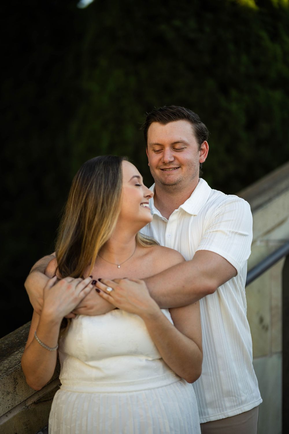 Man embracing woman from behind, both smiling, outdoors near stone steps.
